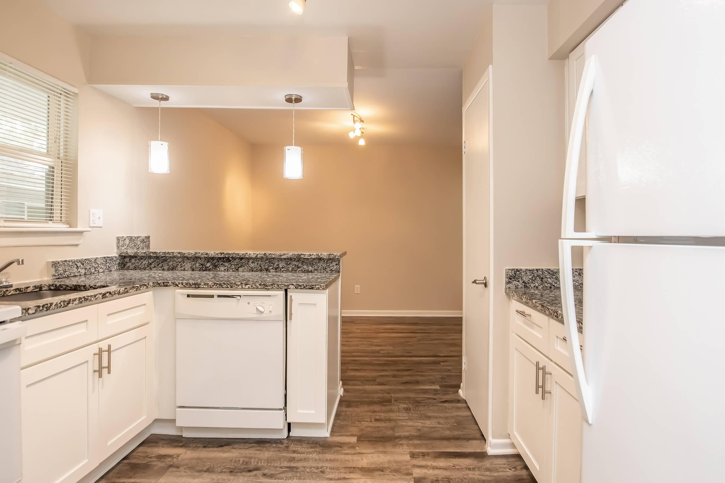 Modern kitchen featuring white cabinetry, granite countertops, a dishwasher, and a white refrigerator. Bright lighting from pendant and recessed fixtures highlights the clean design, while large windows allow natural light. The flooring is a warm wood-like finish, creating an inviting atmosphere.