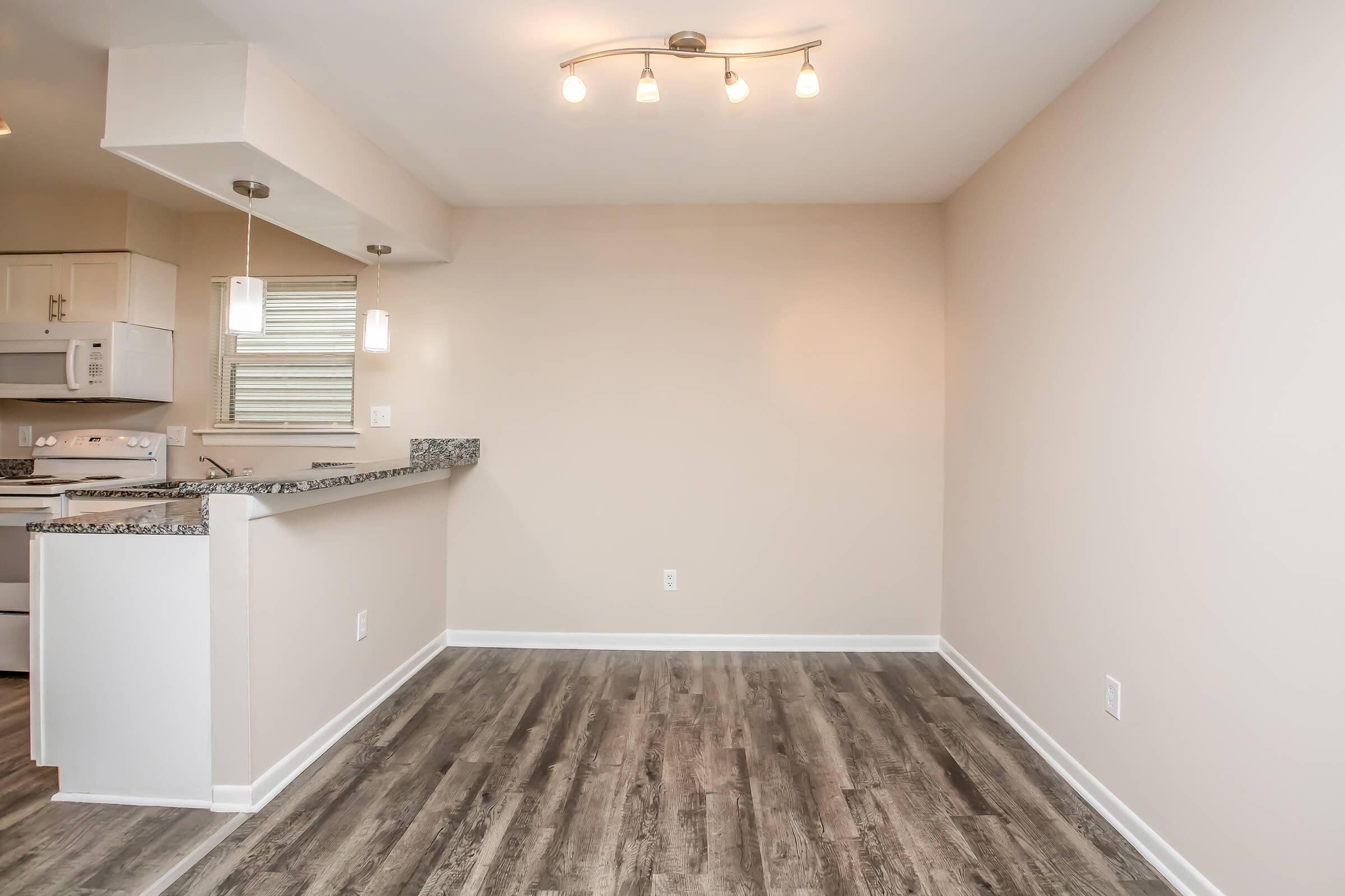 A spacious, empty room with a light-colored wall and wood-like flooring. There is a bar counter with two pendant lights above, and a kitchen area in the background with white cabinets and a stove. The overall feel is modern and inviting, suitable for an apartment or condo.