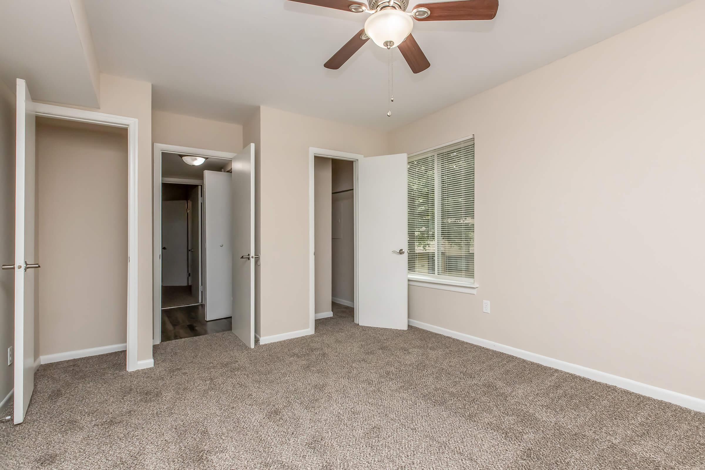 A clean, empty bedroom featuring light beige walls and plush carpet. The room has a ceiling fan, and two open doors leading to closets and a hallway. Natural light streams in through a window, creating a warm, inviting atmosphere.