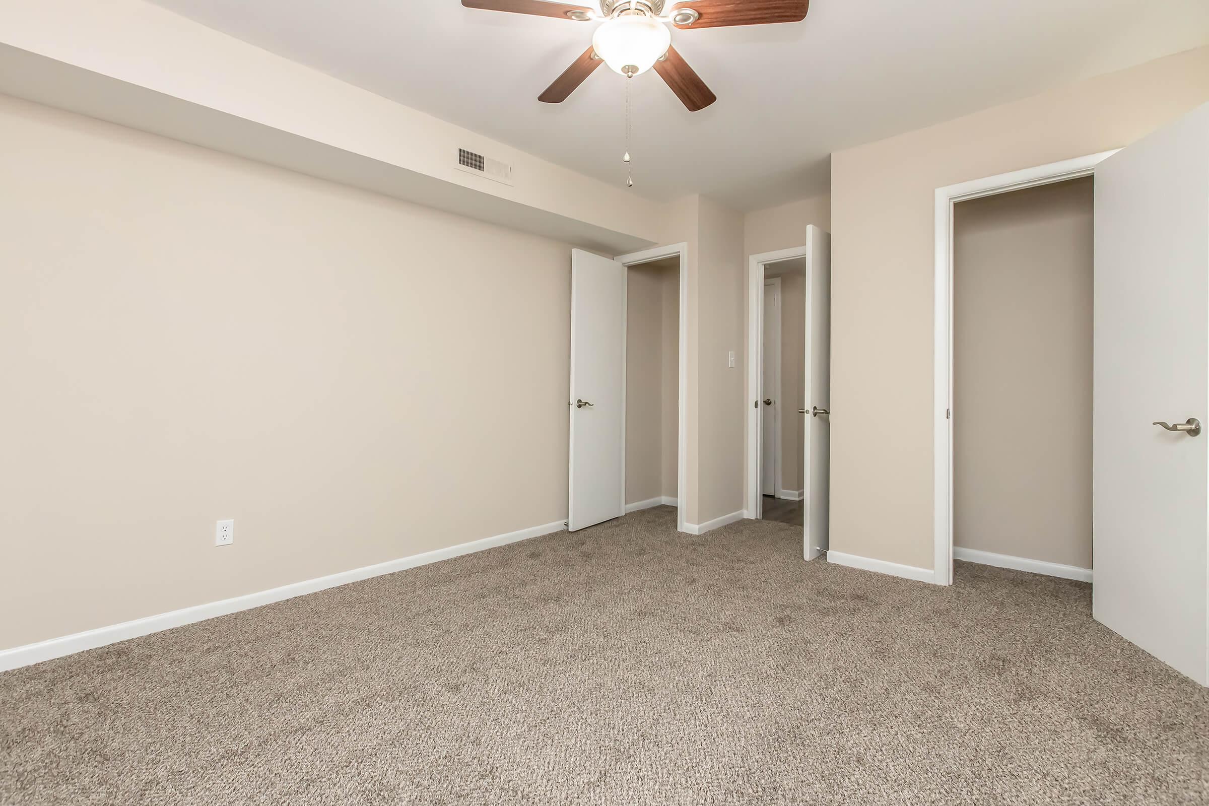 Empty room with beige walls and light brown carpeting. A ceiling fan with a light fixture is visible. There are two open doors on the right leading to closets or other rooms, while one closed door is on the left side of the image. Natural light is absent, creating a neutral and inviting atmosphere.