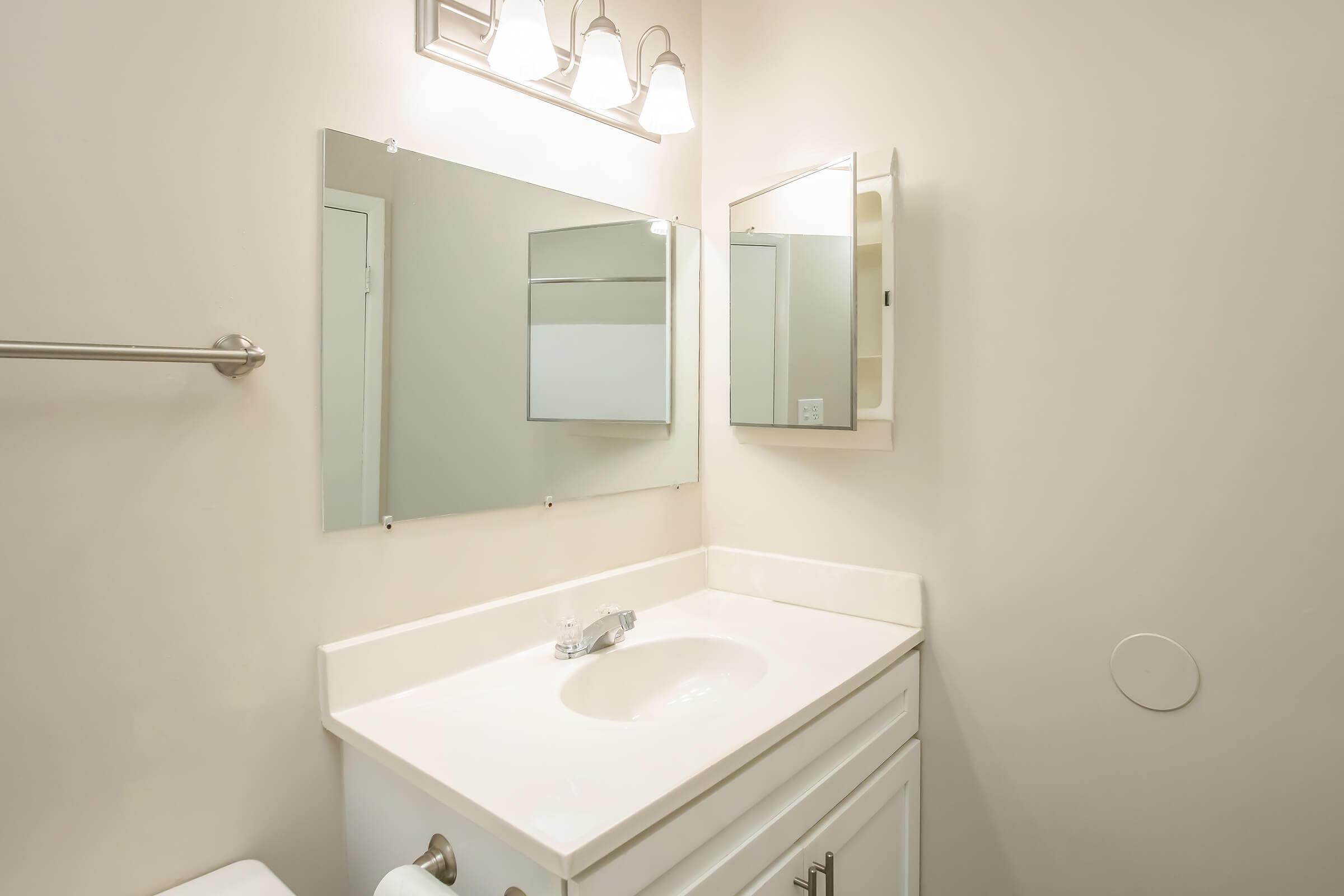 A modern bathroom with a white vanity featuring a sink, a large mirror above the sink, and a light fixture with three bulbs. The walls are painted in a light color, and there is a towel bar on the left. Additional mirror cabinetry is visible, enhancing the space's functionality.