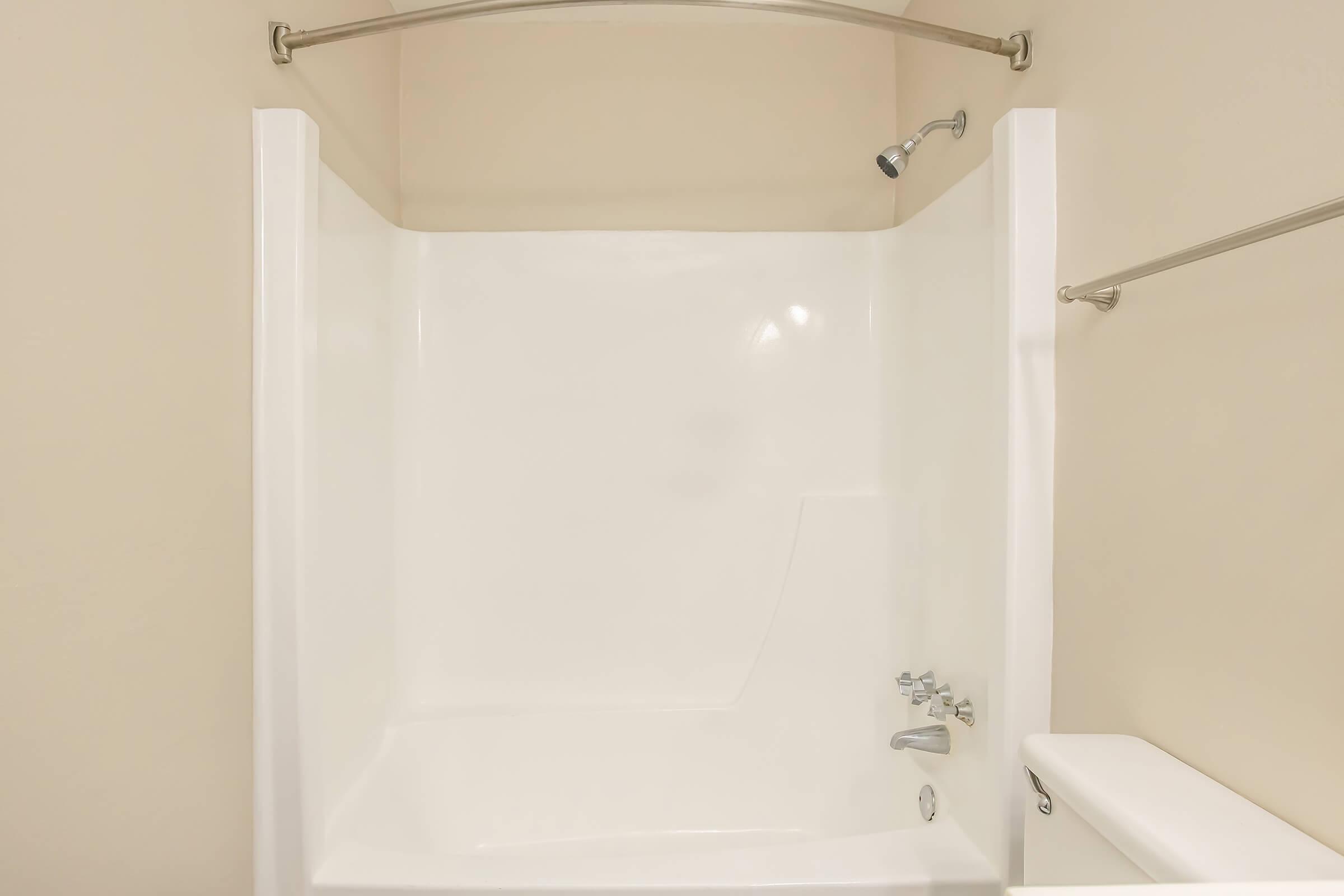 A clean, modern shower stall with a curved shower rod and showerhead. The walls are smooth and white, with a simple, minimalist design. A small ledge is seen next to the faucet controls, and the overall aesthetic is bright and uncluttered, suggesting a fresh, well-maintained bathroom space.