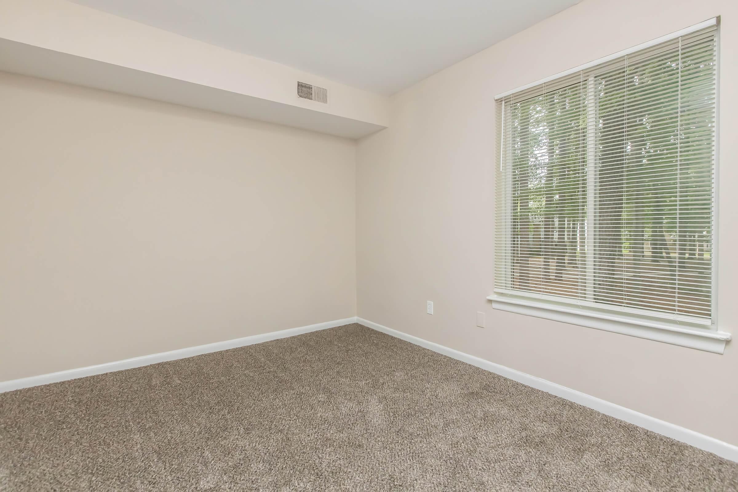 A well-lit, empty room with light beige walls and a large window featuring white blinds. The floor is covered in plush, brown carpet, and there is a small ledge below the window. The overall atmosphere is clean and inviting, with natural light streaming in.