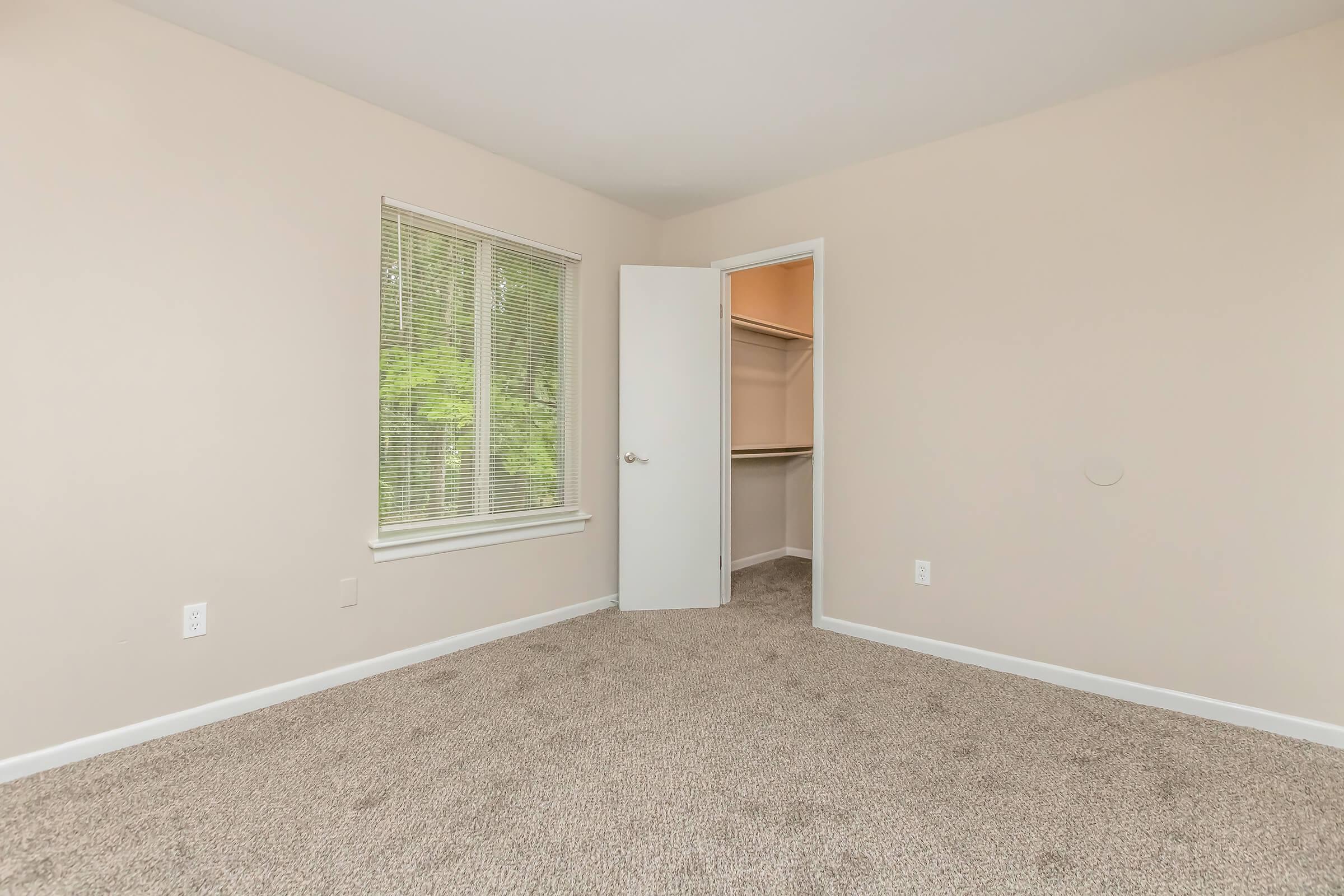 Empty room with light beige walls and a carpeted floor. A window on the left allows natural light to enter, and a door on the right leads to a closet space. The closet has an open door, revealing a simple shelving unit inside. Overall, the room has a clean and minimalist appearance.