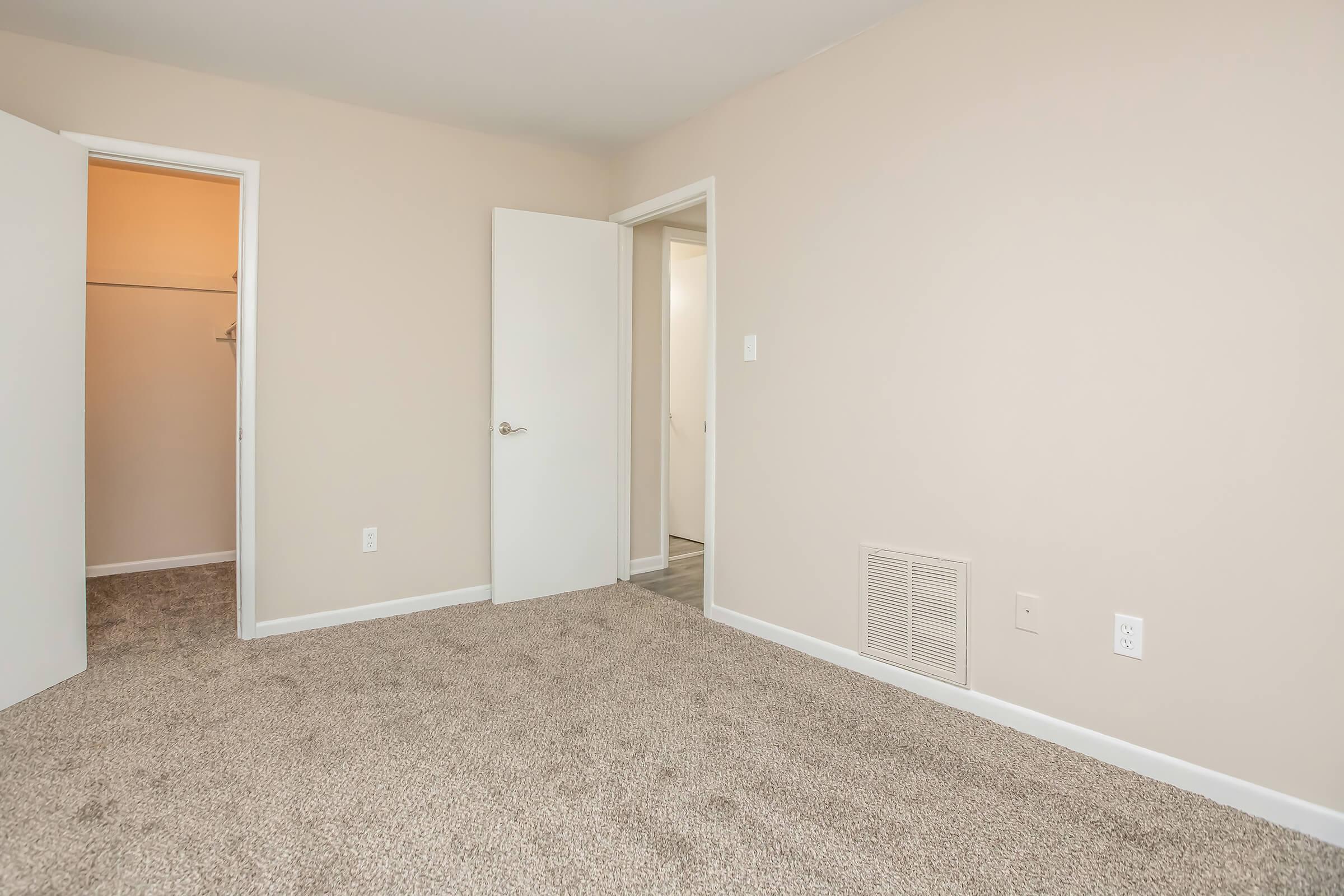 Empty interior of a room with beige walls and carpeted flooring. There are two doors on the left leading to a closet and another room, and a vent near the floor on the right. The overall space is well-lit, with soft lighting visible from the doorways.