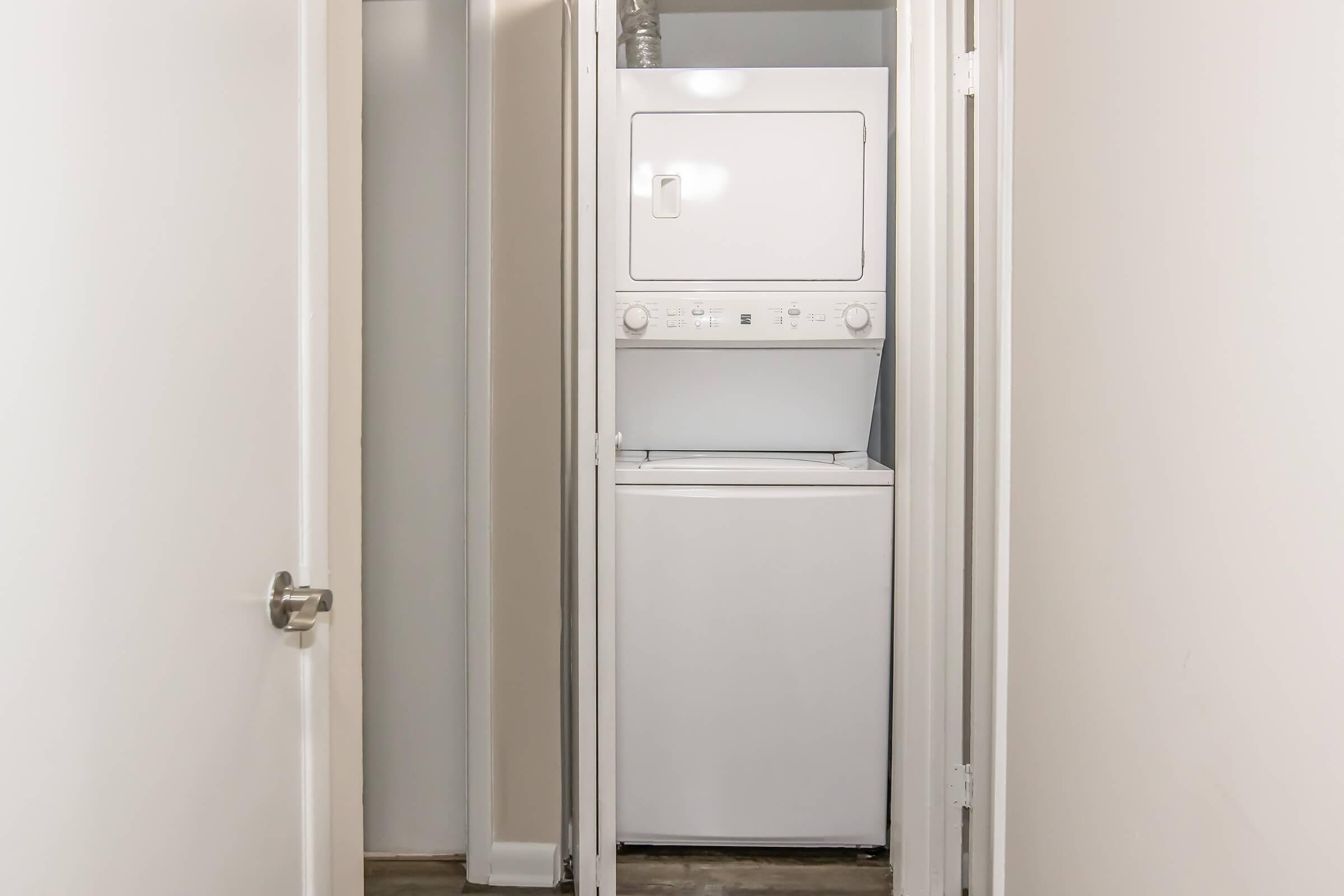 A compact laundry area featuring a stacked washer and dryer unit, positioned between two white doors in a narrow hallway. The walls are painted in neutral tones, and the flooring is a simple, light-colored material.