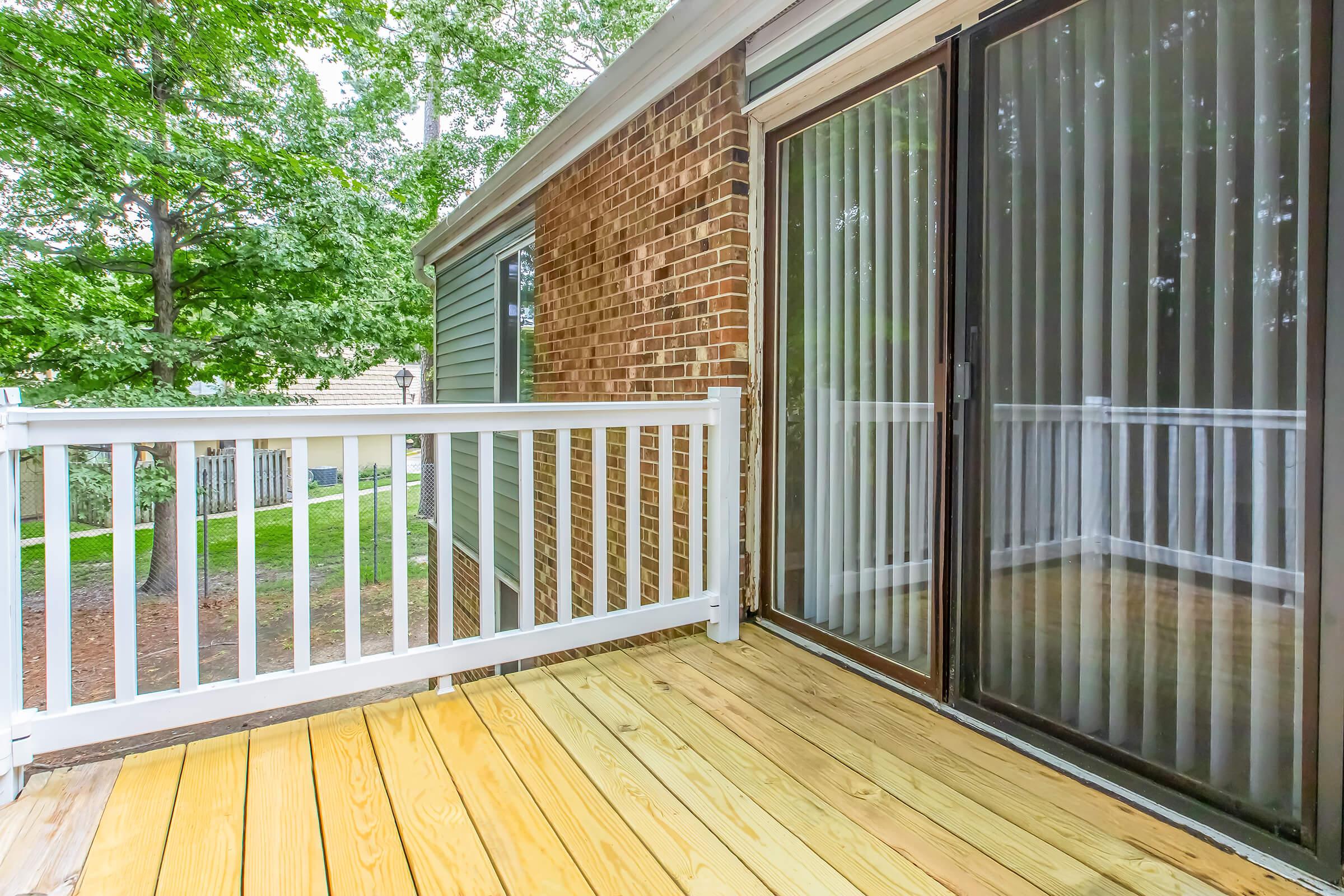 A wooden deck area with a white railing, leading to sliding glass doors surrounded by brick and green siding. Lush green trees are visible in the background, suggesting an outdoor space that is inviting and well-maintained.
