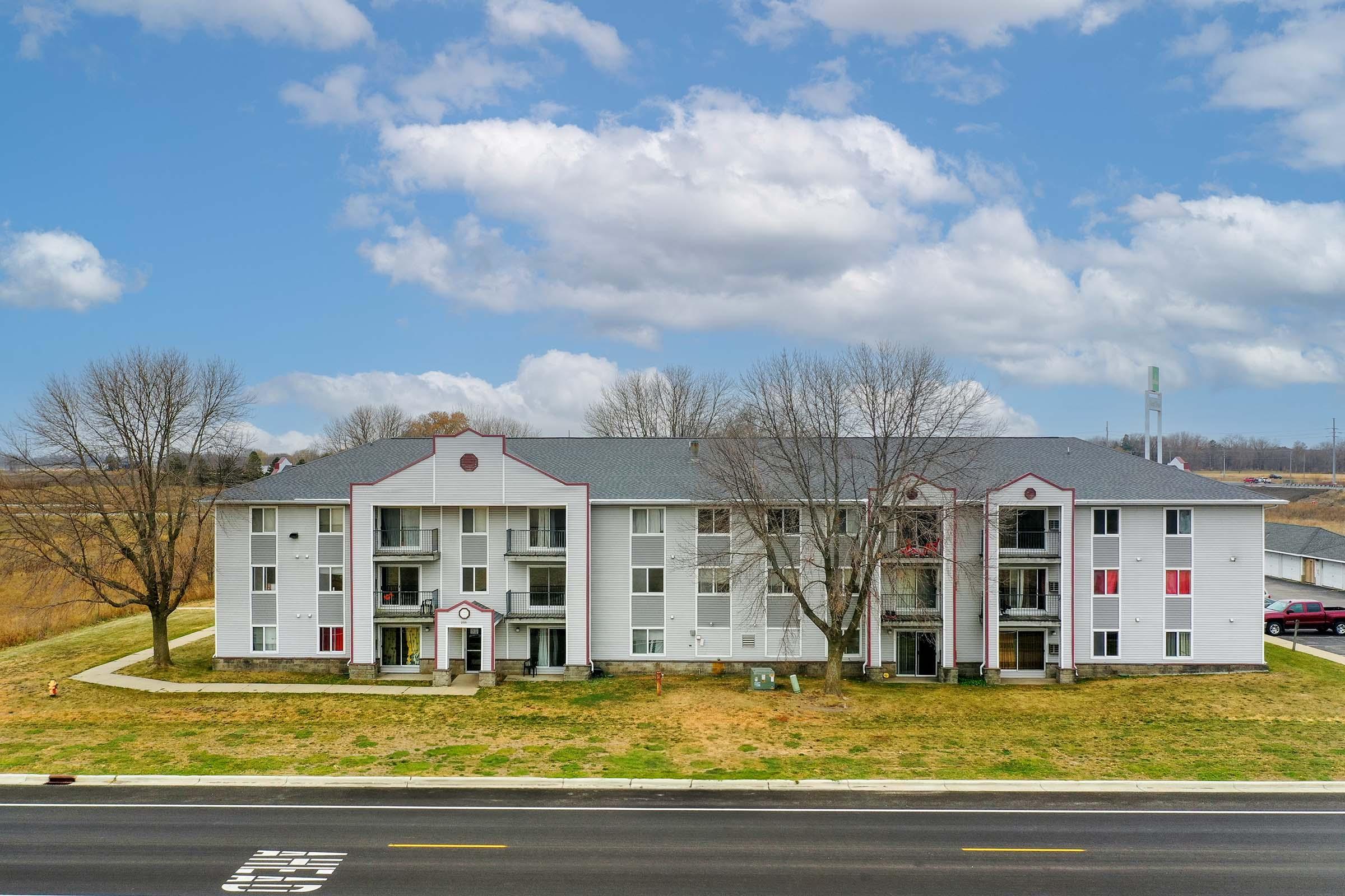 A three-story apartment building with a white exterior and red accents, set against a cloudy blue sky. Trees in front are bare, and a grassy area surrounds the building. The scene is located near a road, with fields visible in the background.