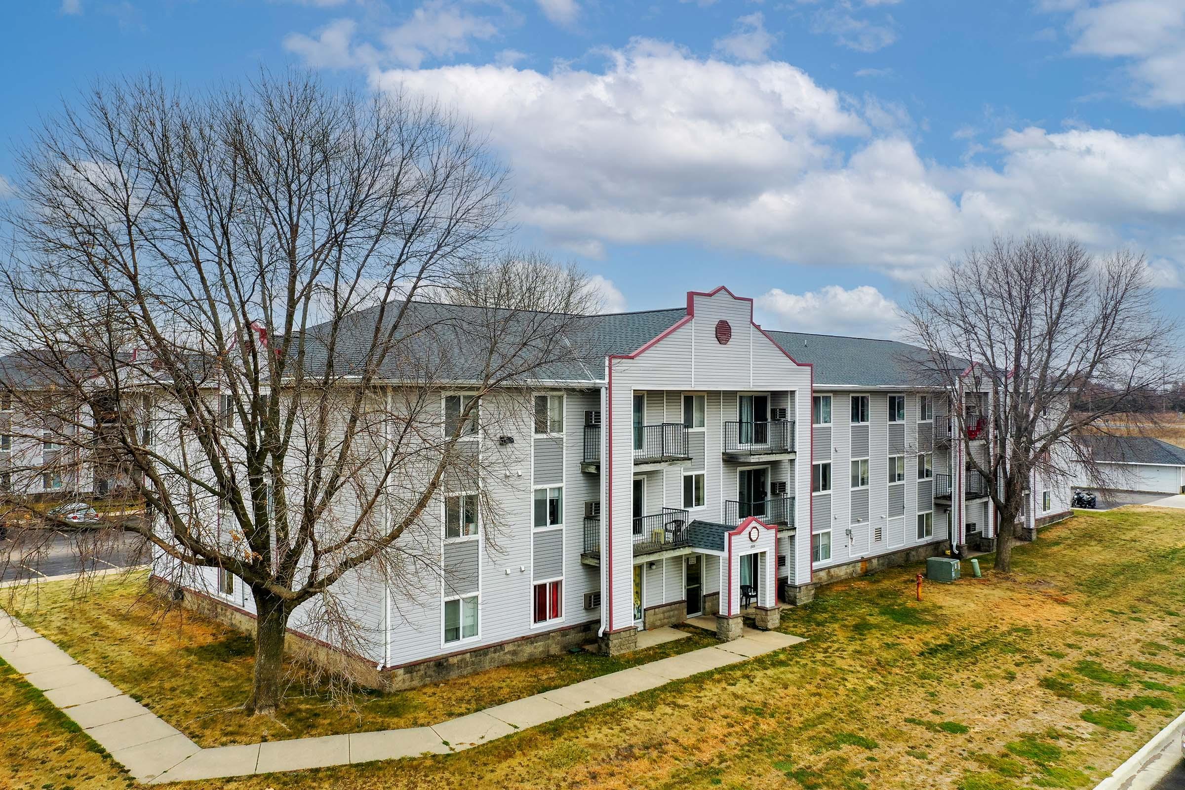 A three-story apartment building with a white exterior, red accents, and multiple balconies. The surrounding area features a grassy lawn and bare trees, under a partially cloudy sky. A pathway leads from the street to the building's entrance.