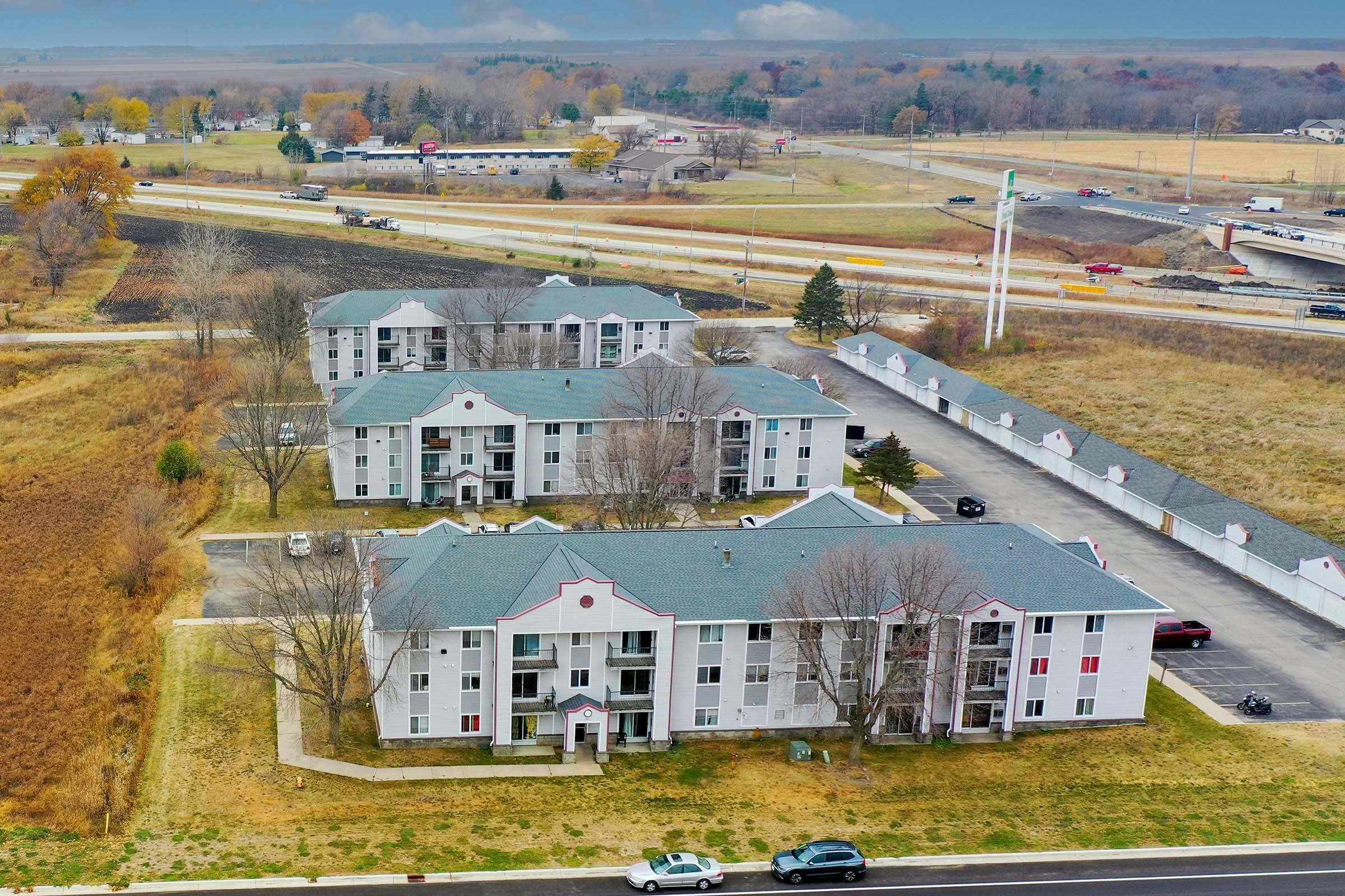 Aerial view of a residential complex with multiple buildings, gray roofs, and surrounding grassy areas. In the background, a highway and open fields can be seen. The scene is set under a partly cloudy sky, indicating an expansive landscape. Several parked cars are visible near the buildings.