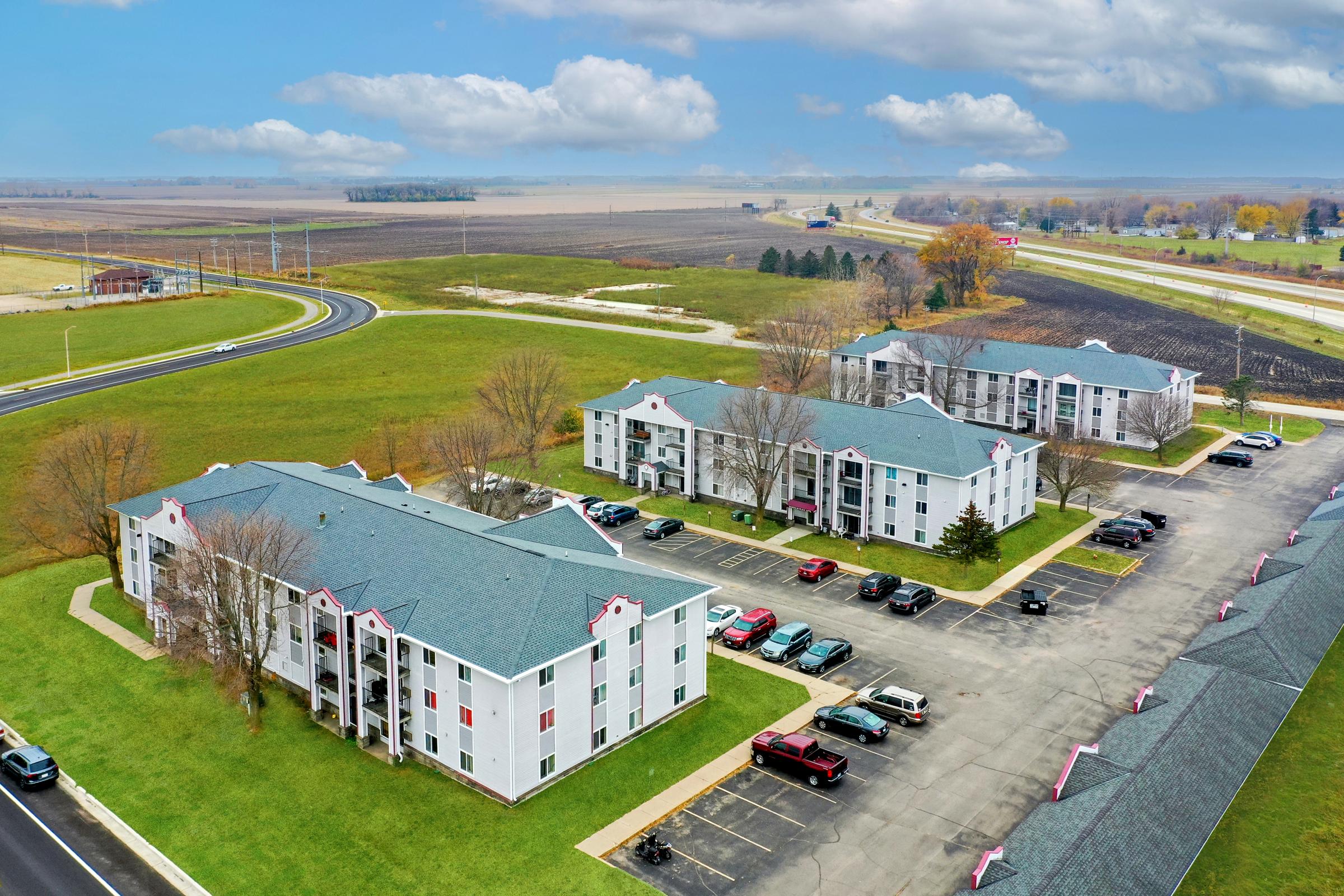 Aerial view of a multi-building complex, featuring two three-story residential buildings with white exteriors and dark roofs. Surrounding green lawns and a parking lot filled with cars are visible, along with an open field and road in the background under a cloudy sky.