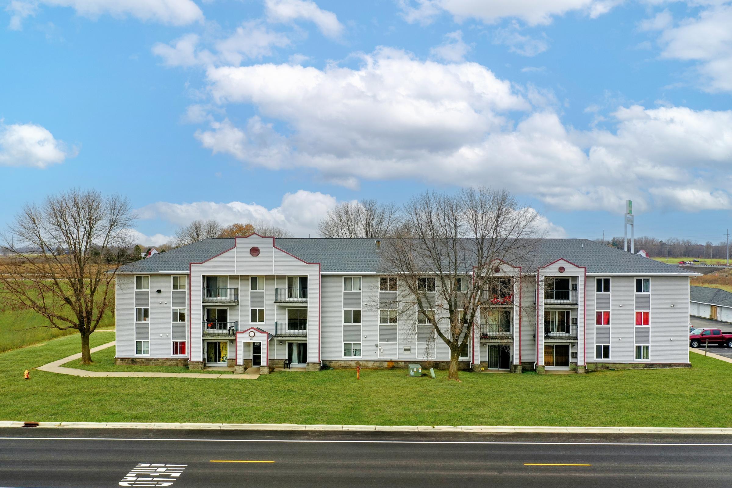 A three-story residential building with a white and gray exterior, featuring multiple balconies and red accents. The surrounding area includes a grassy lawn and a road in the foreground, with a clear blue sky and clouds overhead.