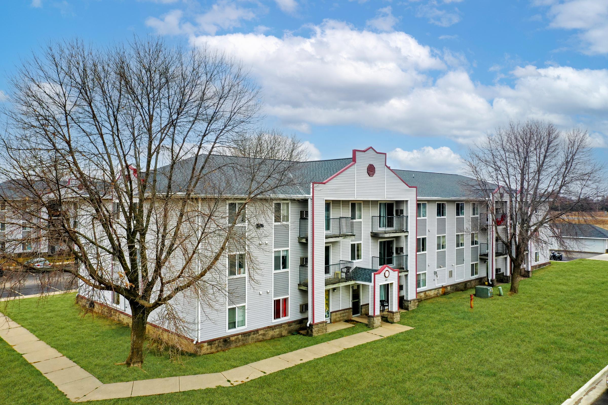 A three-story apartment building with a gray roof and white siding, featuring a decorative facade. Surrounding the building is a grassy area with two bare trees and a concrete walkway leading to the entrance. The sky is partly cloudy, adding a serene backdrop to the scene.