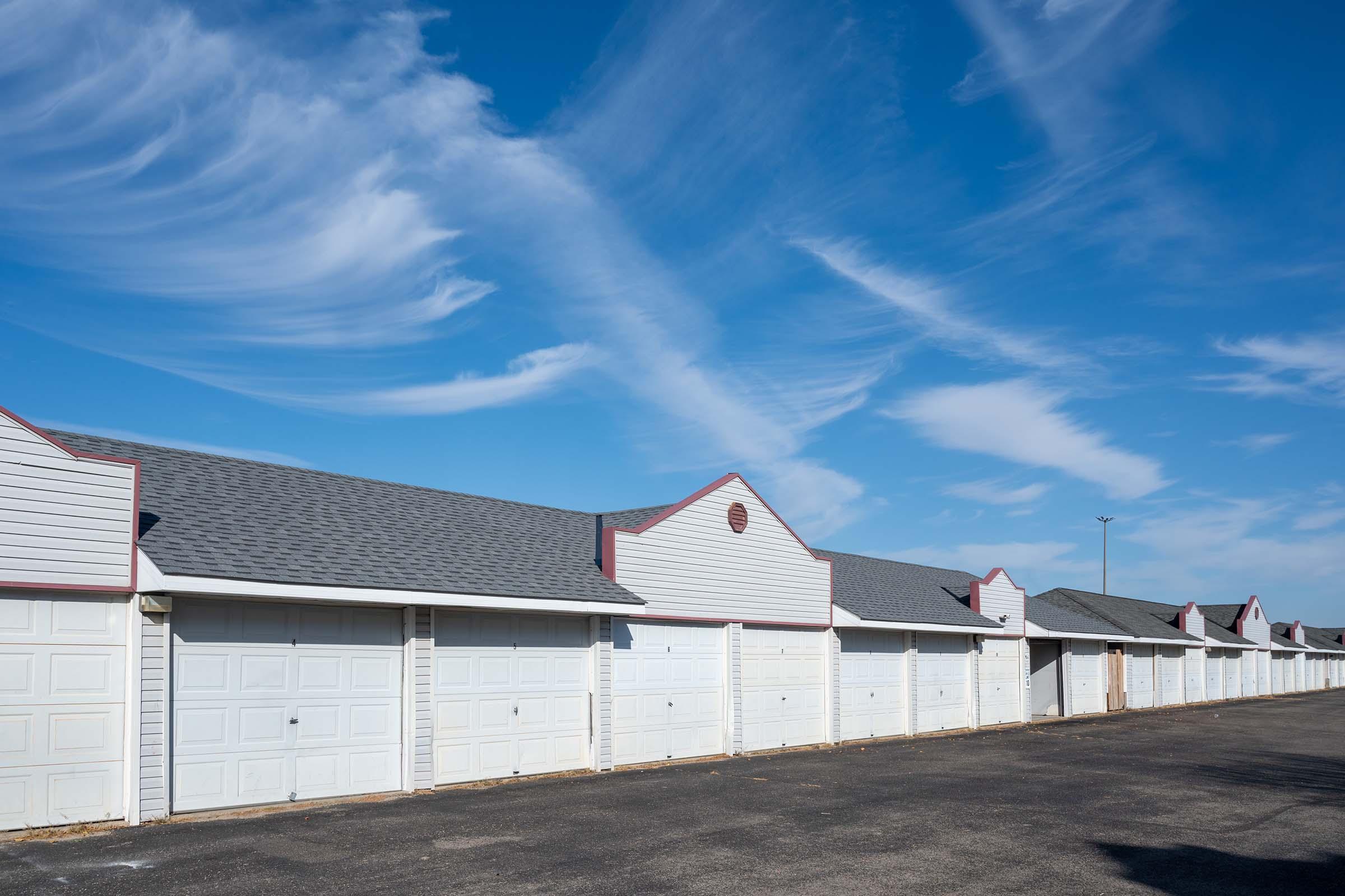 A row of closed garage doors lined up against a clear blue sky with wispy clouds. The garages have white exteriors and peaked roofs, creating a uniform appearance along the pavement. The scene is calm and conveys an organized, suburban environment.