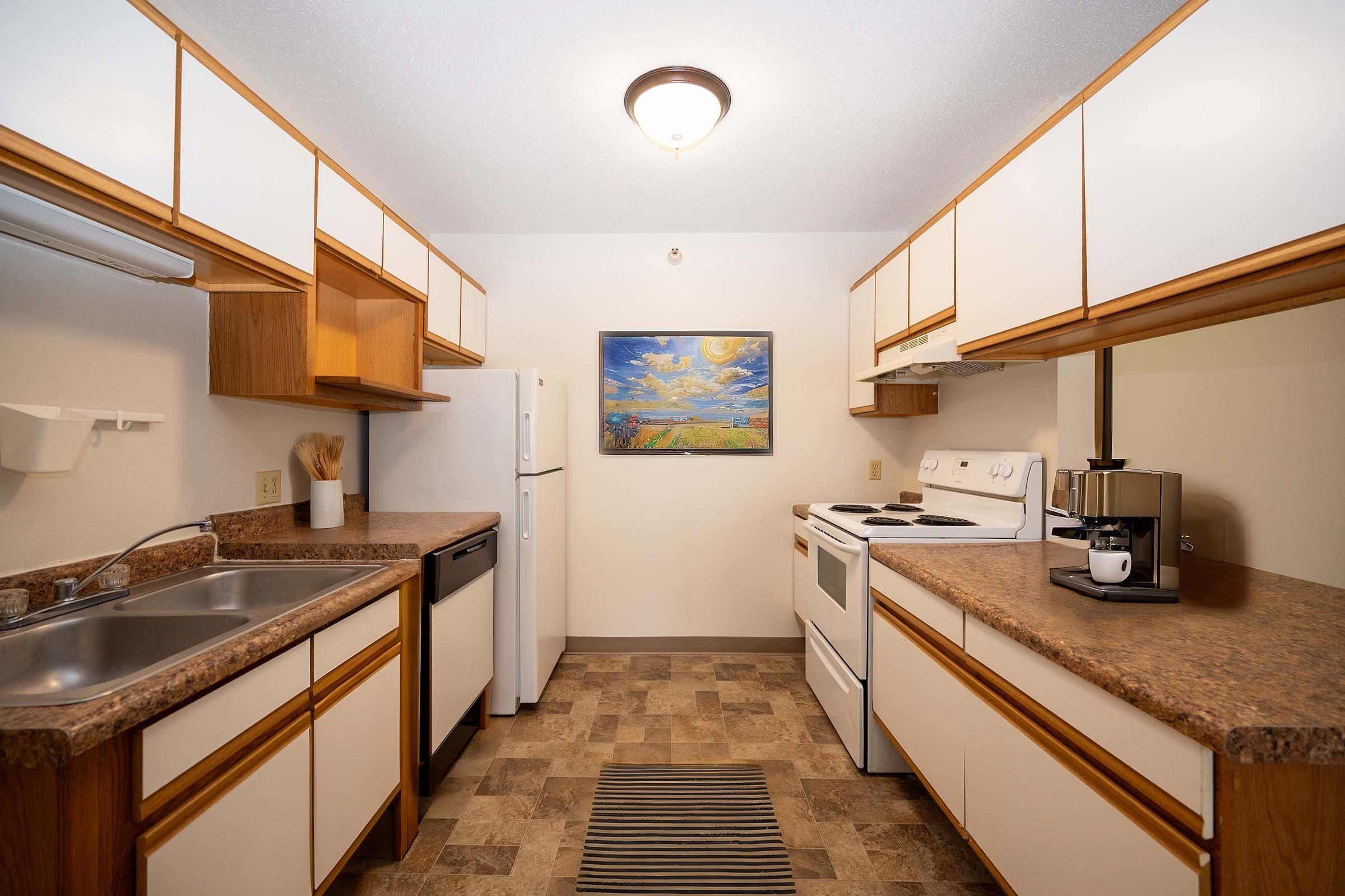 A clean, modern kitchen featuring a white refrigerator, stove, and sink with wooden cabinets and countertops. A colorful painting hangs on the wall, and a coffee maker sits on the counter. The floor is covered with a striped rug, creating a cozy atmosphere.