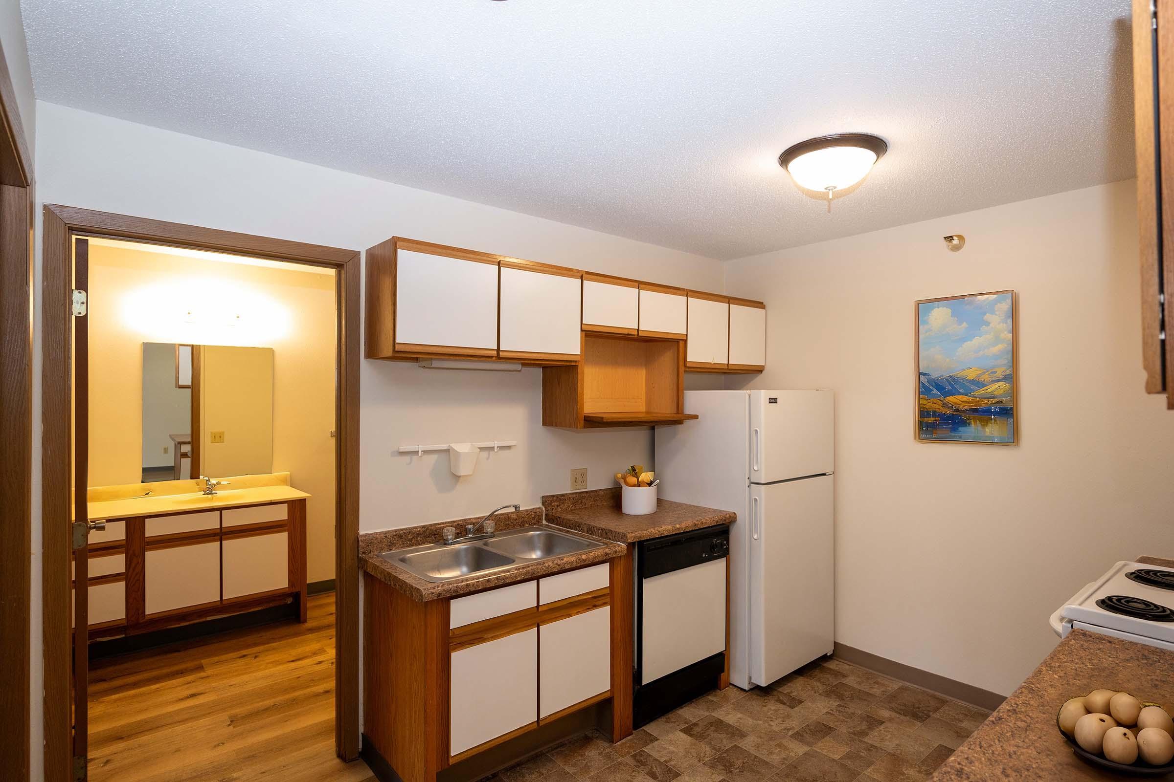 A kitchen featuring wooden cabinets, a double sink, a refrigerator, and a stove. The room has beige walls and a light fixture on the ceiling. A doorway leads to a bathroom with a mirror and vanity area, while a painting of mountains decorates one wall. The floor is covered with light-colored tiles.