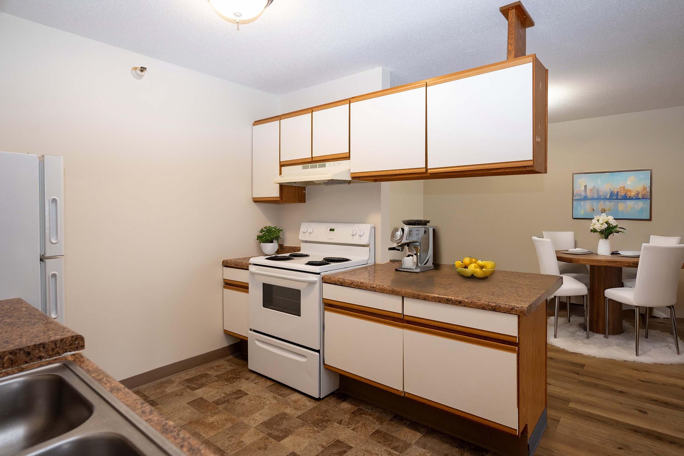 A modern kitchen featuring a white stove, refrigerator, and coffee maker. The countertops are brown with a smooth finish. There's a bowl of yellow fruit on the countertop, and a dining area with a round table and white chairs in the background. Soft lighting creates a warm atmosphere.