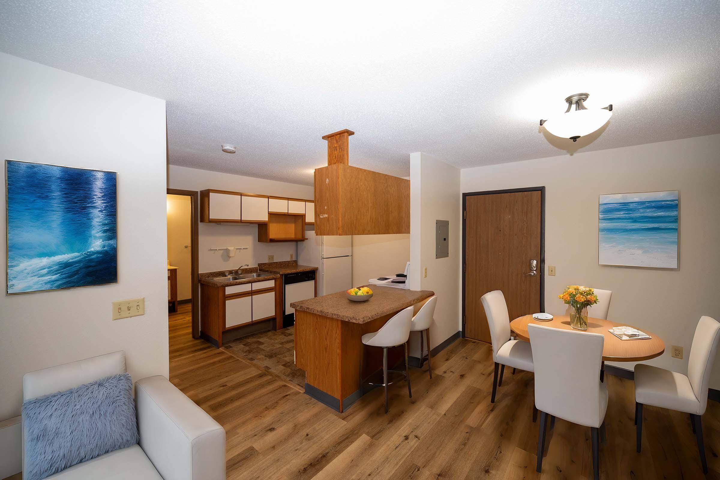A cozy apartment interior featuring a modern kitchen with a dining area. There’s a small table with four white chairs, a beige sofa, and artwork depicting ocean scenes on the walls. The kitchen has wooden cabinets, and a light fixture illuminates the space. The flooring is wooden, adding warmth to the atmosphere.