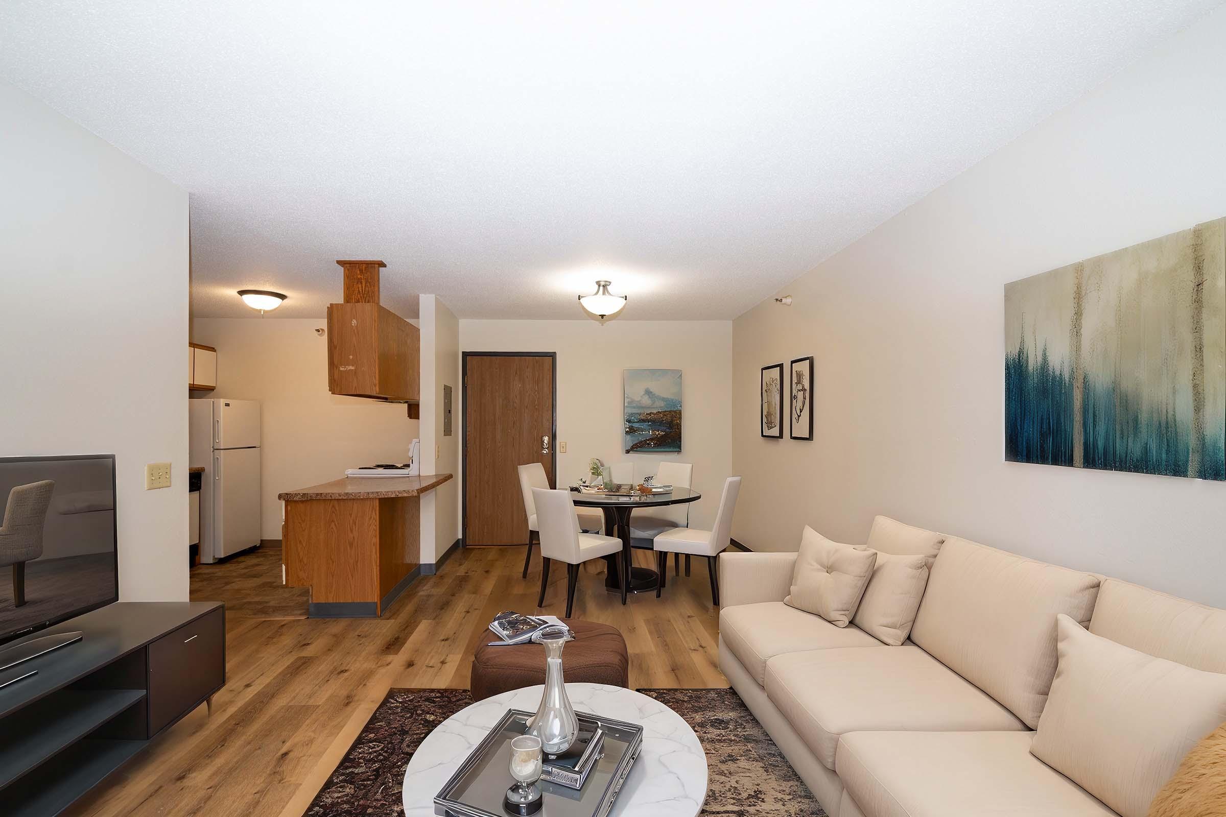 A modern living area featuring a light-colored sofa with pillows, a round dining table with chairs, and a kitchen area in the background. The hardwood floor adds warmth, and there are decorative wall art pieces. A coffee table sits in front of the sofa, enhancing the inviting atmosphere of the space.