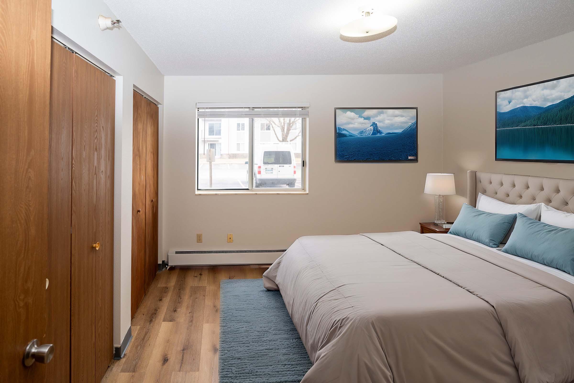 A cozy bedroom featuring a queen-sized bed with light blue pillows and a beige headboard. There are two wooden doors on the left, a lamp on a nightstand beside the bed, and a large window with a view of a parked vehicle outside. A landscape painting hangs on the wall above the bed.