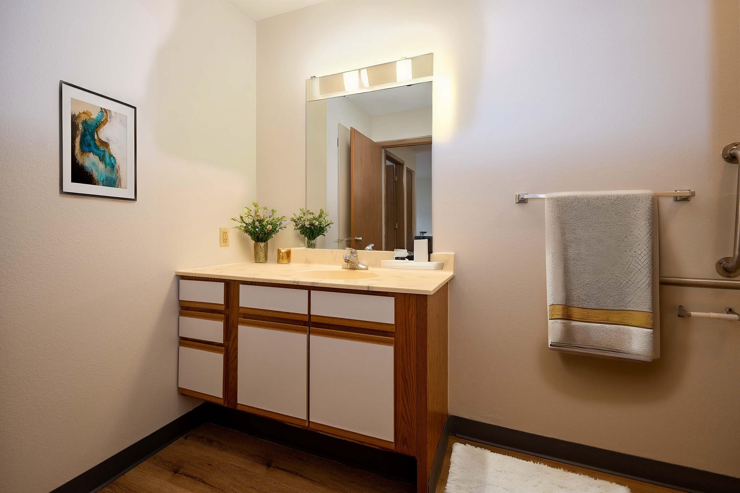 A modern bathroom featuring a light-colored vanity with wooden accents, a sink, and a mirror above. There are framed artwork on the wall, a towel hanging on a rack, and a small arrangement of flowers on the counter. The floor is covered with a soft white rug.