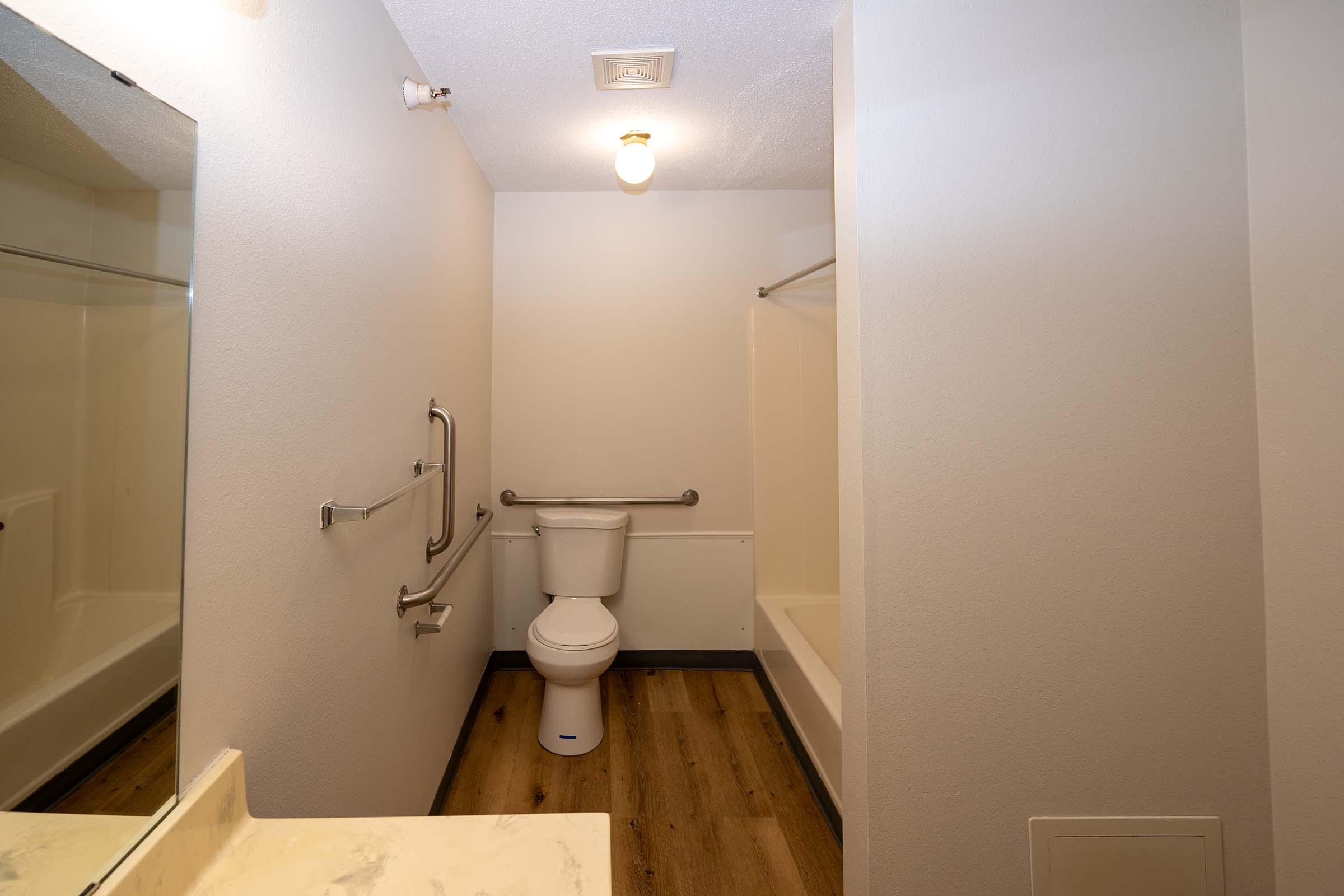 A clean, modern bathroom with a beige and white color scheme. Features include a toilet, a bathtub with a shower curtain, a wall-mounted grab bar, and a mirror above the vanity. The floor is a light wood texture, and there's a ceiling light providing illumination.