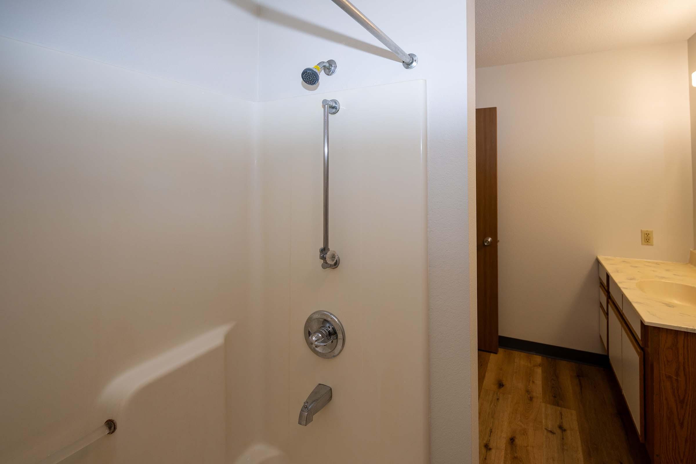 A clean, modern bathroom featuring a white shower stall with a handheld showerhead and grab bar. Light wood flooring contrasts with the white walls and shower. A simple vanity counter with cabinets is visible in the background, alongside a closed wooden door.