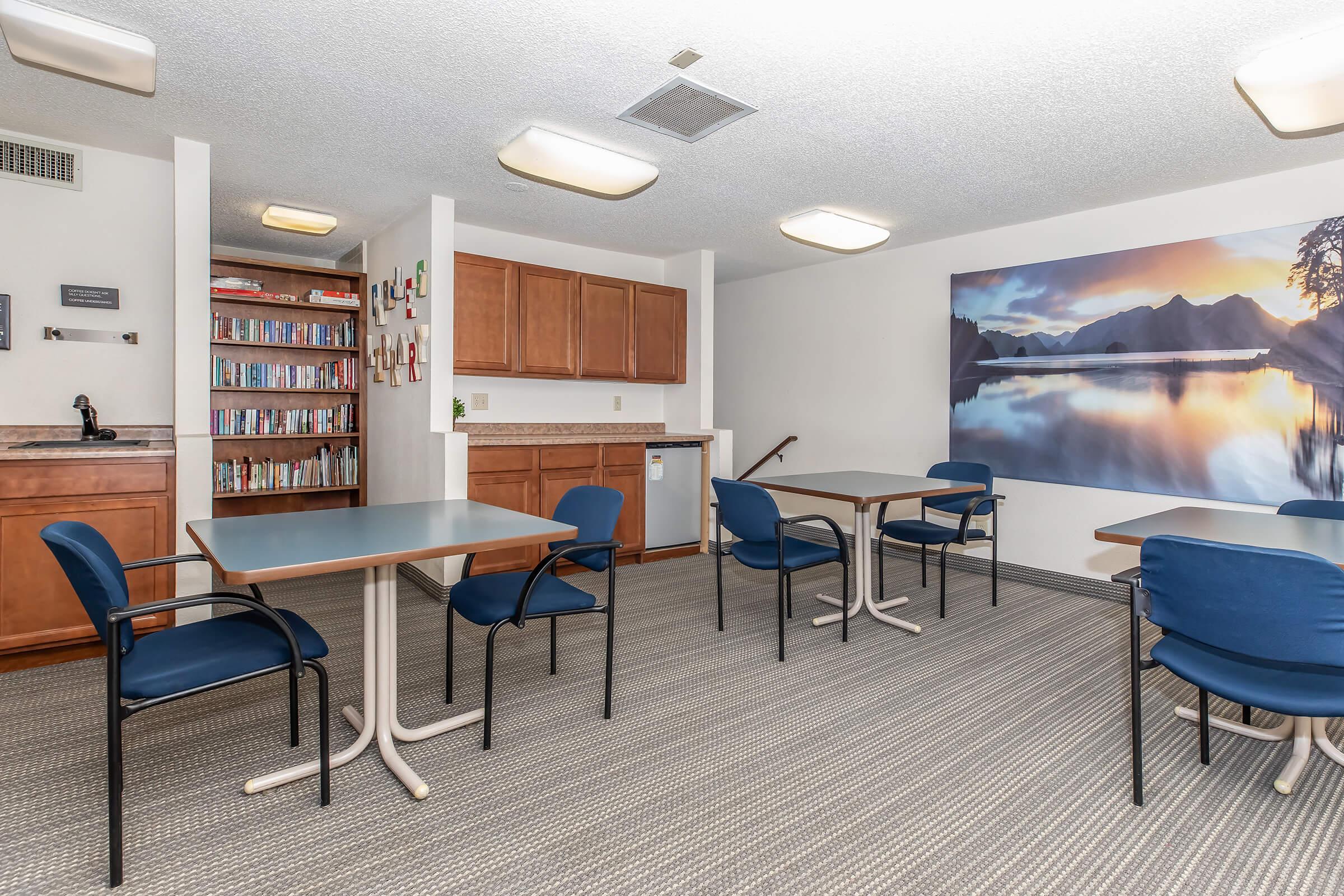 A spacious common room featuring several blue chairs and tables arranged for use. The area includes a kitchenette with wooden cabinets, a bookshelf filled with various books, and a large mural of a serene landscape with mountains and water, enhancing the inviting atmosphere.