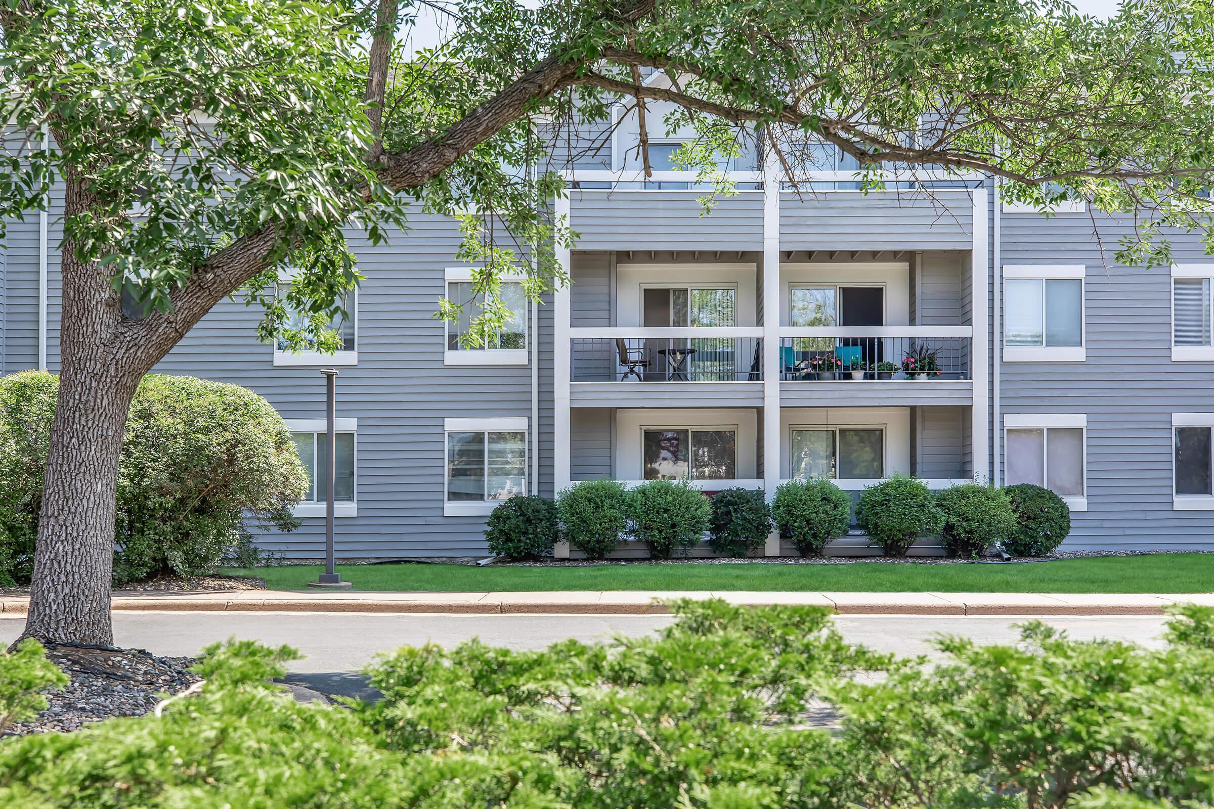 A modern apartment building with multiple balconies, framed by lush greenery. The exterior features a muted gray facade and large windows, while decorative shrubs line the front. A tree provides shade, creating a pleasant outdoor environment. Ideal for residential living.