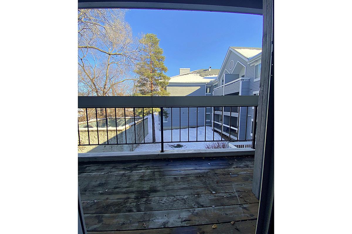 View from a balcony overlooking a snow-covered yard and adjacent buildings. The scene features a wooden railing, a clear blue sky, and signs of winter with snow on the ground. Bright light highlights the peaceful outdoor setting.