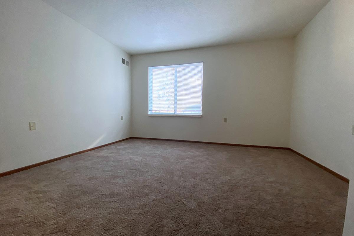 A minimally furnished room with light brown carpet and plain white walls. There is a single window with white blinds allowing natural light to enter. The space is empty, creating a spacious and uncluttered atmosphere.