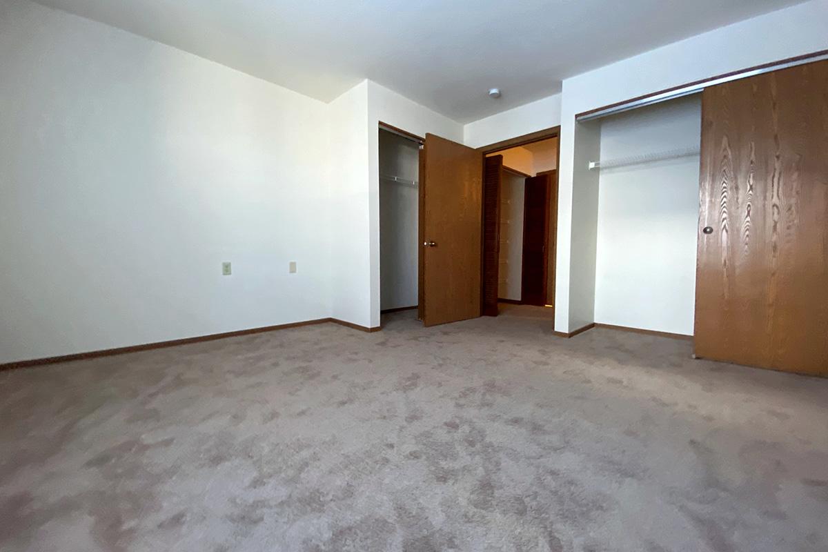 A vacant room with light-colored walls and beige carpet. There are two wooden doors standing partially open, leading to closets. The room is well-lit, with natural light coming from an unseen source, creating a spacious and empty atmosphere.