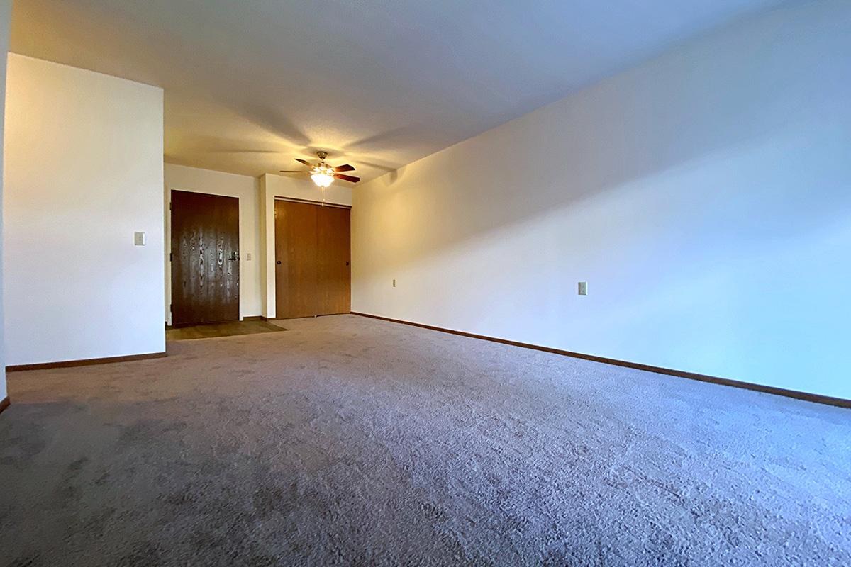 A spacious room with beige carpet and white walls, featuring a ceiling fan. There are two closed wooden doors, one on the left and one on the right. The lighting is soft, and the overall ambiance is bright and inviting. The room appears empty, providing a versatile space for various uses.