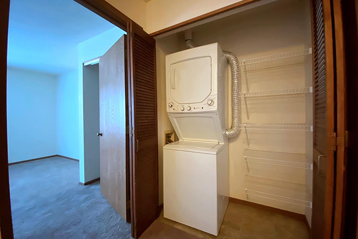 A compact laundry area featuring a stacked washer and dryer unit. Adjacent to it is a wire shelf for storage. The space is well-lit and has a door leading to another room, with a simple carpeted flooring in the area.