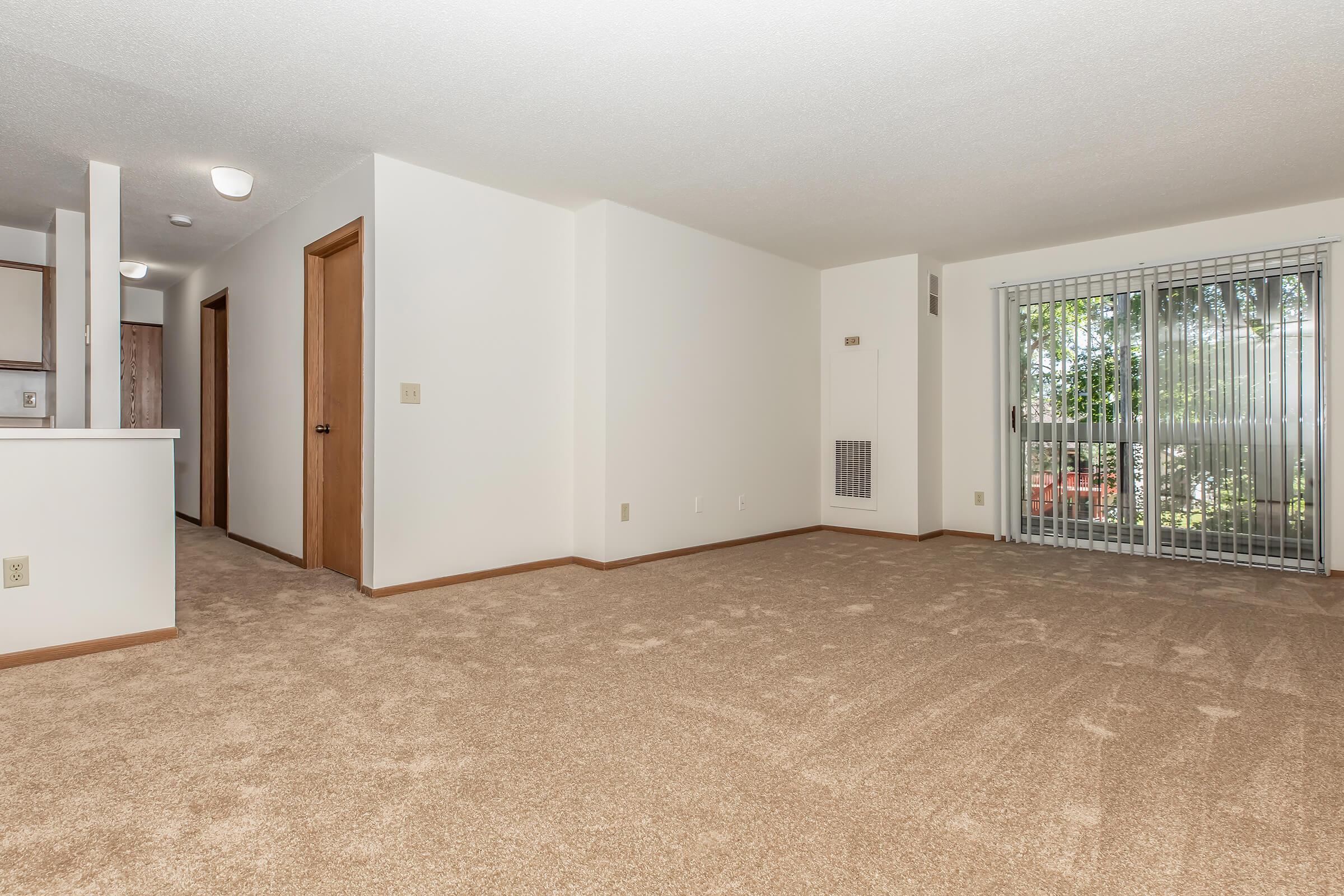 A spacious, empty living room featuring light-colored walls and carpet. The room has two doors leading to other areas, and a large window with blinds offers natural light and a view of an outdoor space. The overall atmosphere is bright and inviting, ideal for personalizing.