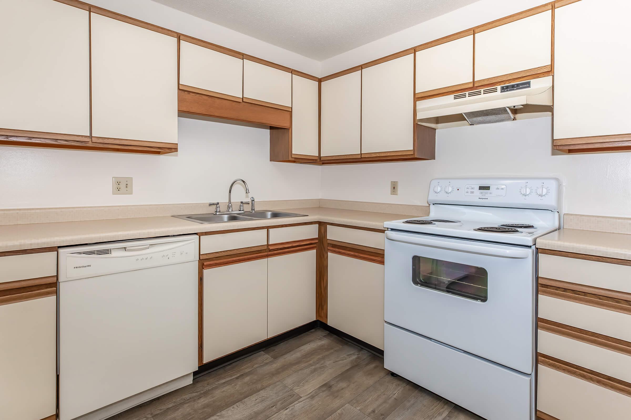 A modern kitchen featuring light-colored cabinets with wooden accents, a white stove, and a dishwasher. The countertop is beige, and a double sink is installed under a wall-mounted cabinet. The floor has a wood-like finish, creating a warm and inviting atmosphere.