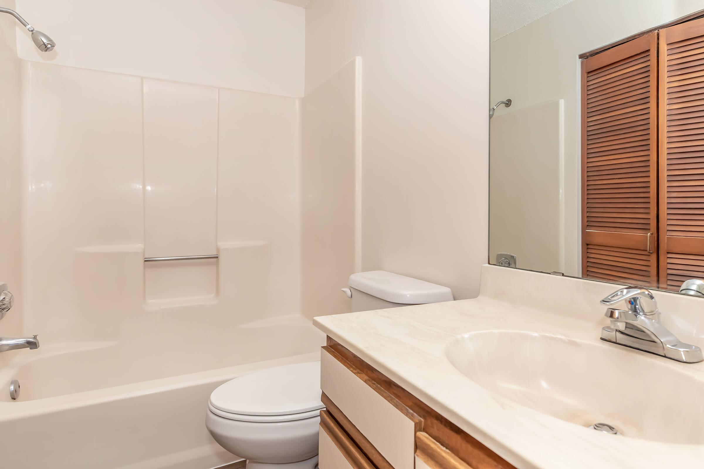 A clean, modern bathroom featuring a bathtub with a shower, a toilet, and a countertop with a sink. The walls are light-colored, and a large mirror reflects the space. There are wooden cabinets beneath the sink, and the room has a neutral, uncluttered appearance.