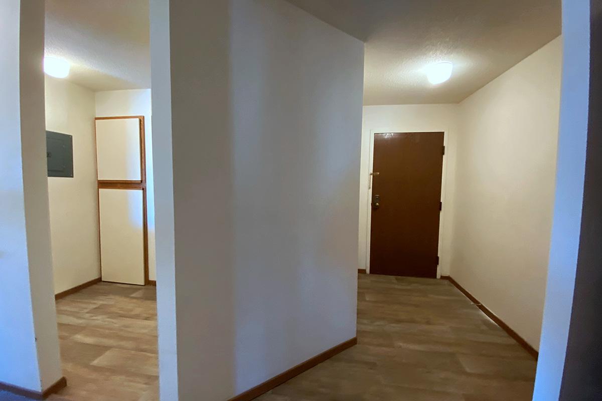 Interior view of a hallway featuring two walls and a closed wooden door. There are light fixtures on the ceiling and a section of a wall with a mailbox or utility panel. The flooring is tiled in a light color, and the walls are painted a neutral shade, contributing to a bright and open atmosphere.