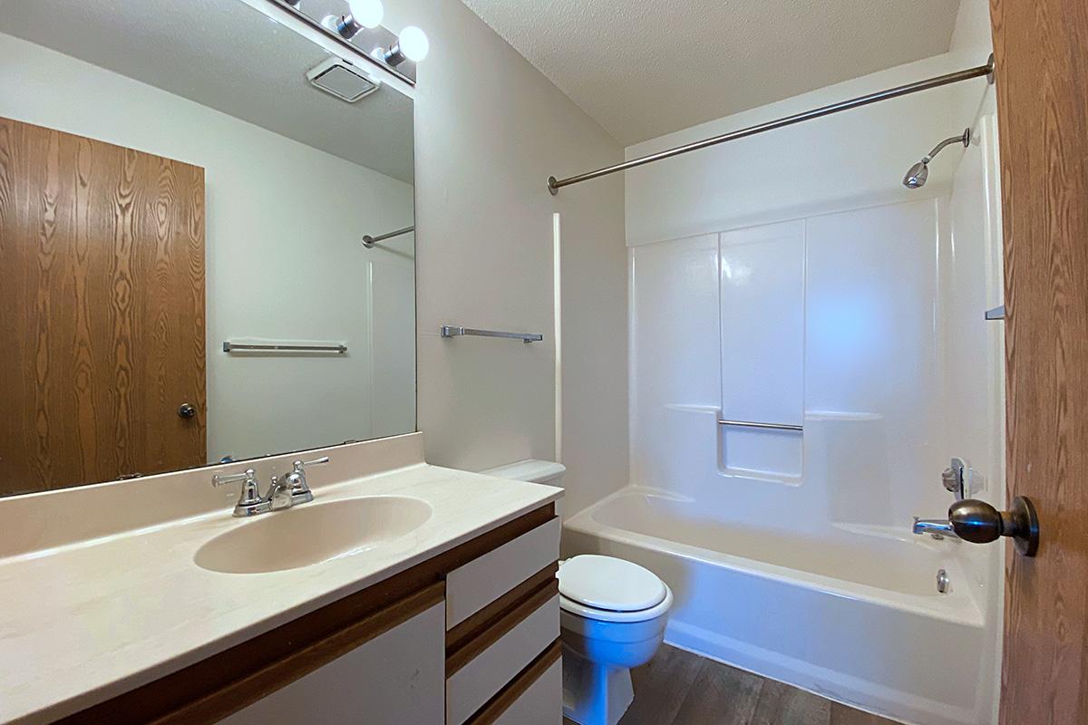 A bright and clean bathroom featuring a white tub and shower combination, a beige countertop with sink, wooden cabinets, a round toilet, and a large mirror above the sink. The walls are light-colored, and there's a wooden door visible, adding a touch of warmth to the space.