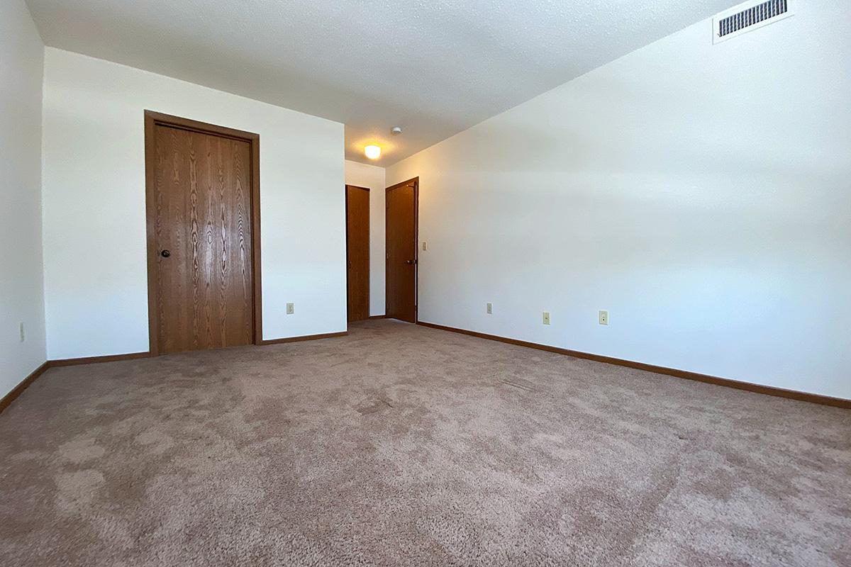 A spacious, empty room with light beige carpet flooring and plain white walls. Two wooden doors on the left lead to adjacent rooms, and a ceiling light fixture is visible. The room is well-lit with natural light, creating a clean and minimalistic atmosphere.