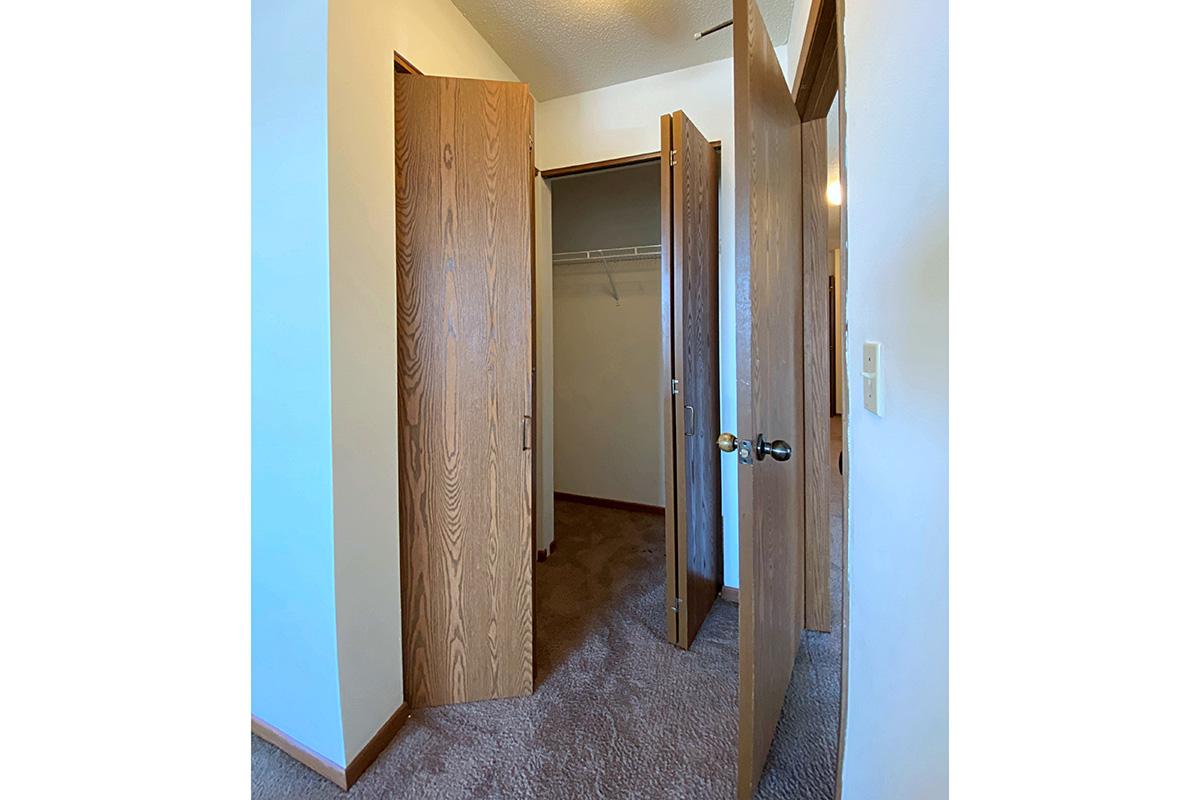 A partially open closet featuring wooden doors, revealing a carpeted floor and a hanging rod inside the closet. The walls are painted in a light color, and there is a light switch visible on the right side of the image.