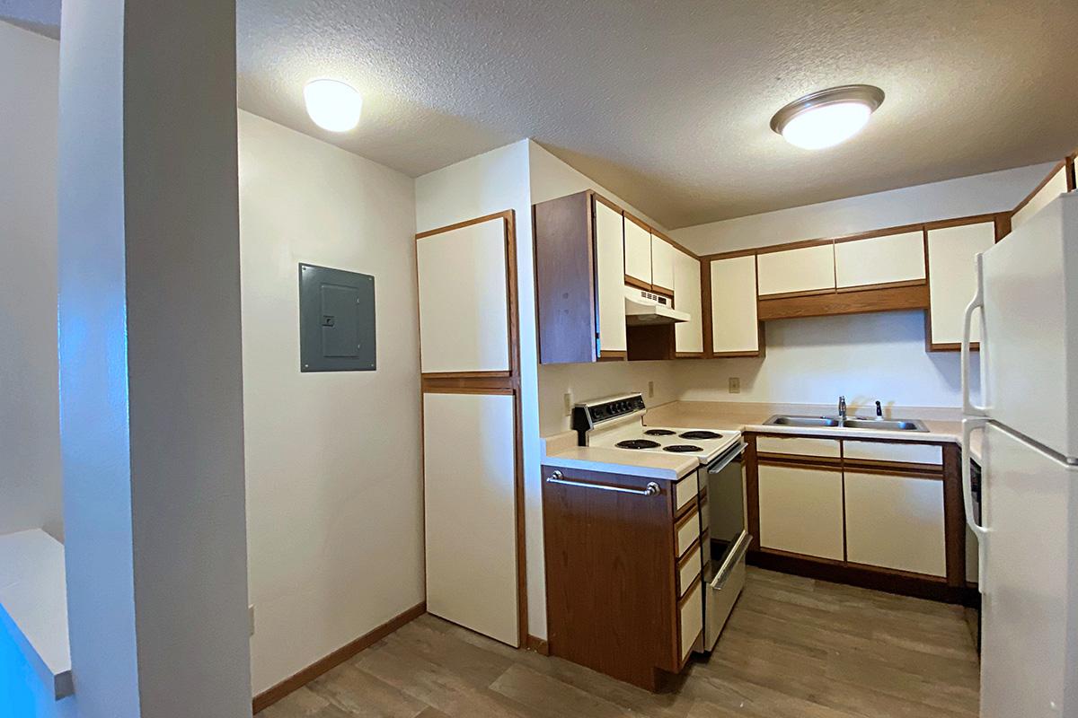 A small, modern kitchen featuring white and wooden cabinetry, a stove and oven combination, a sink with a faucet, and a refrigerator. The room has neutral-colored walls and a light fixture on the ceiling, with a utility panel visible on one wall. The flooring is a light wood finish.