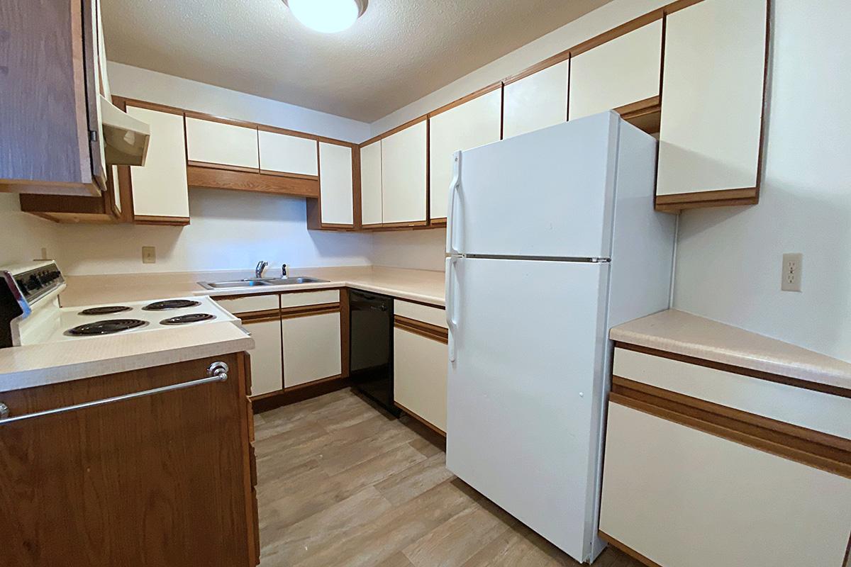 A compact kitchen featuring white cabinets and countertops with wood accents. The space includes a white refrigerator, a sink with a faucet, a stove with four burners, and a dishwasher. The flooring is light-colored laminate, and overhead lighting provides adequate illumination.