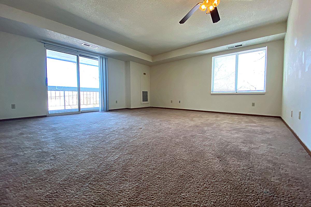 A spacious, empty room with light beige carpet and cream-colored walls. Natural light streams in through a large window on the left, and there is a sliding glass door leading to a balcony. A ceiling fan is visible in the center of the room, contributing to the airy atmosphere.