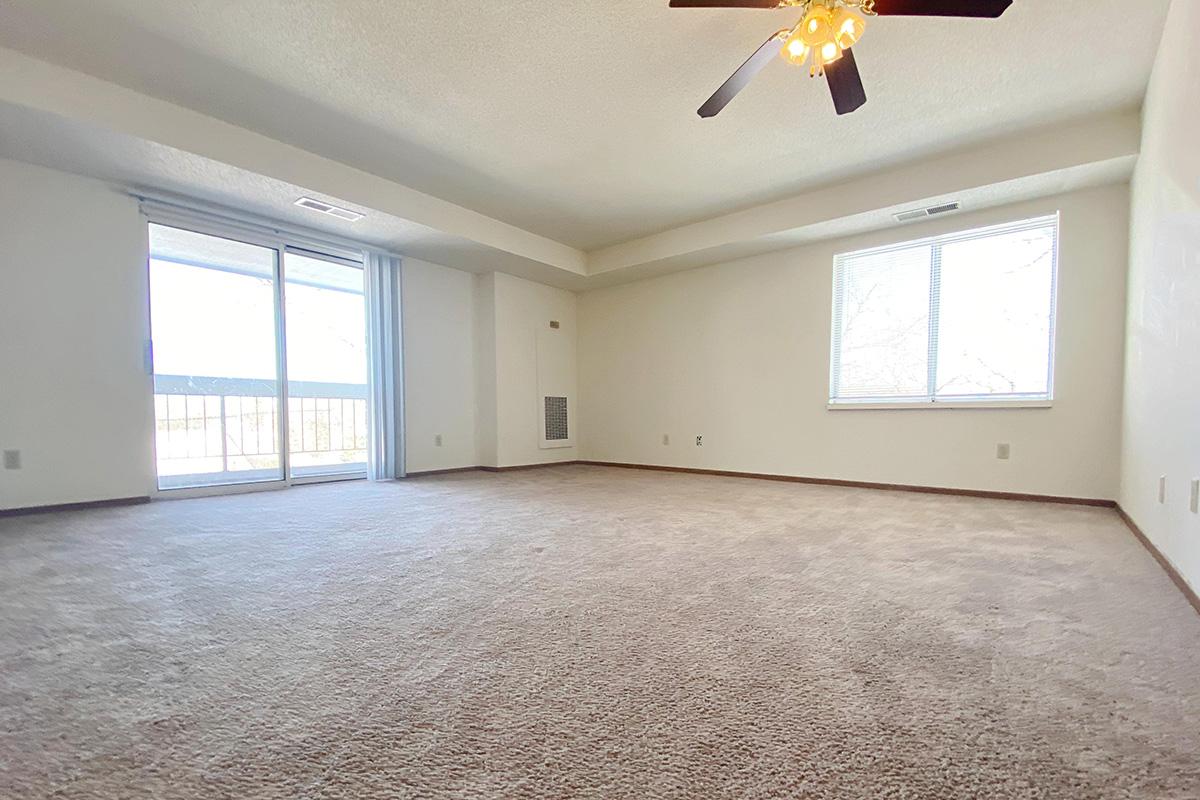 A spacious, empty living room with light beige carpet and neutral-colored walls. A ceiling fan with light is visible. There is a sliding glass door leading to a balcony, and a large window allowing natural light to enter. The room has a clean, open feel, perfect for personalization.