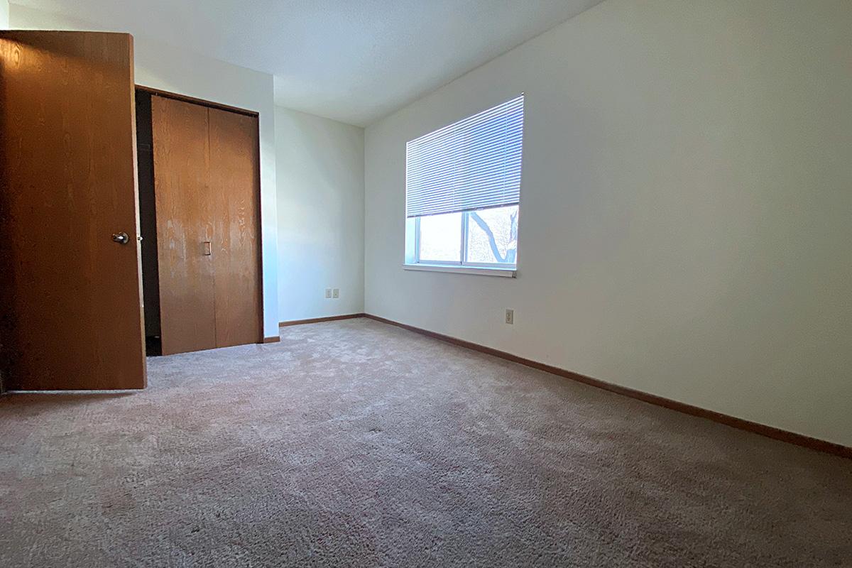 A well-lit, empty room with light-colored walls, a window with closed blinds, and beige carpet. There is a closet with wooden doors partially open on the left side. The space is minimal and undecorated, allowing for various layout possibilities.