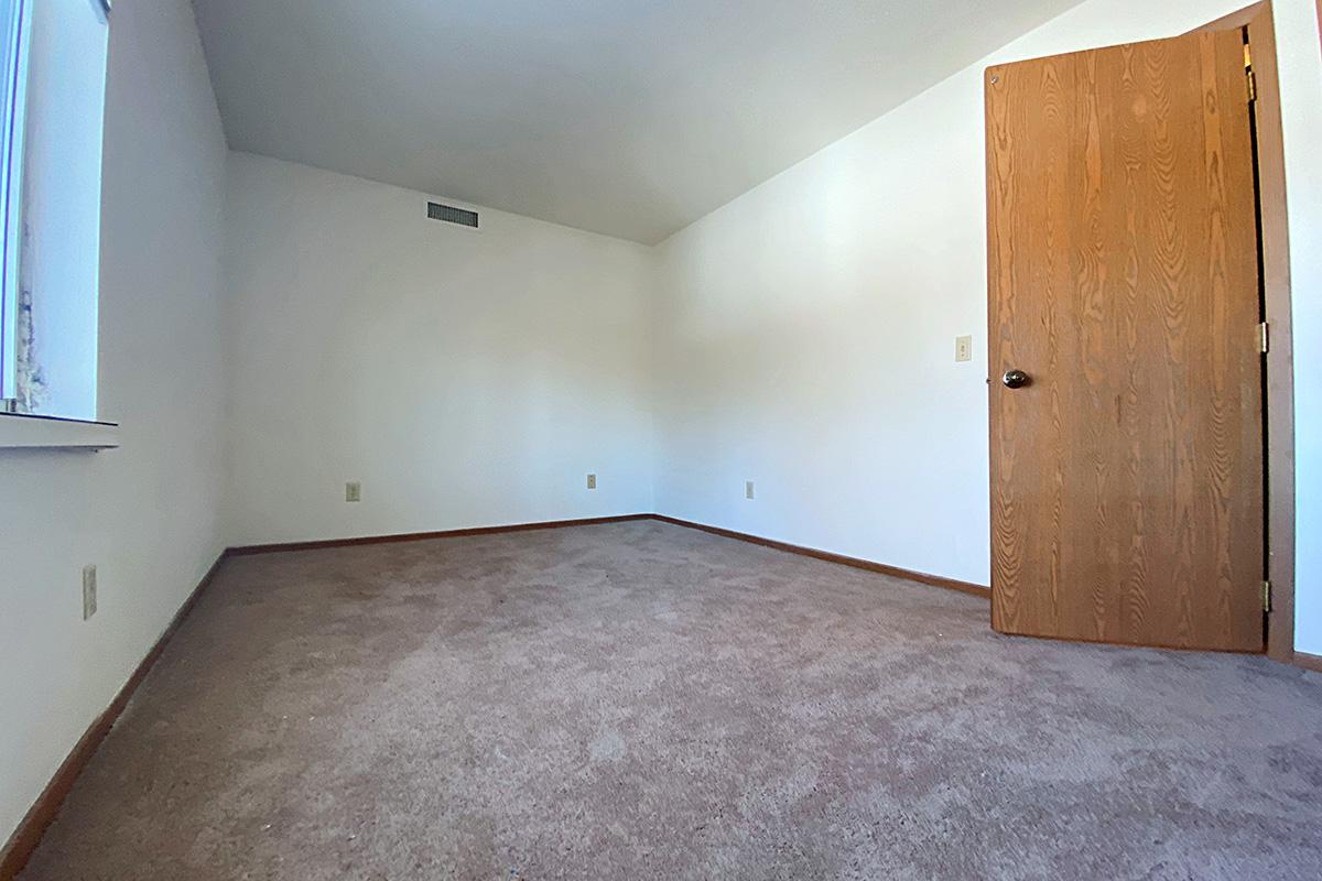 A sparsely furnished empty room with beige carpet and light-colored walls. There is a single wooden door on the right side of the image, and a small window on the left. The ceiling is slanted, creating a spacious appearance. The room is well-lit and appears clean and ready for use.