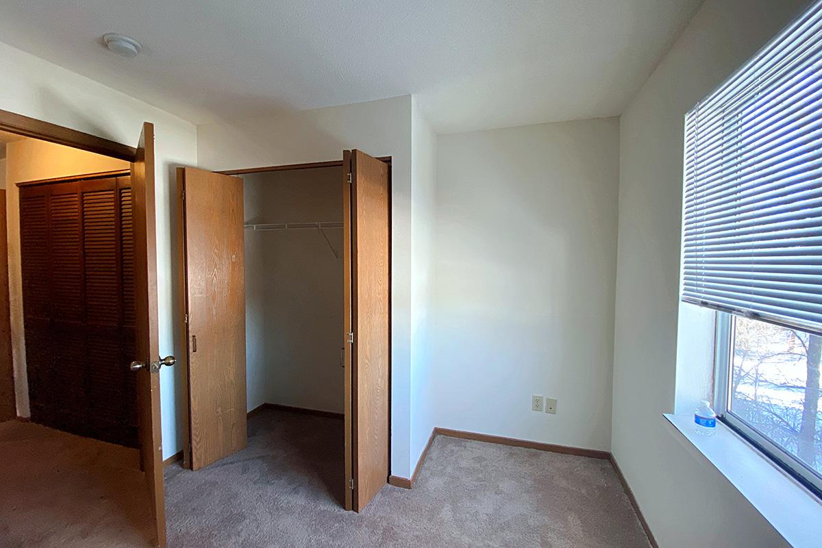 A vacant bedroom with light-colored walls and carpeted floor. One side features an open closet with sliding doors, while the other has a window partially covered by blinds, letting in natural light. The room appears clean and minimalistic, ready for furnishings.