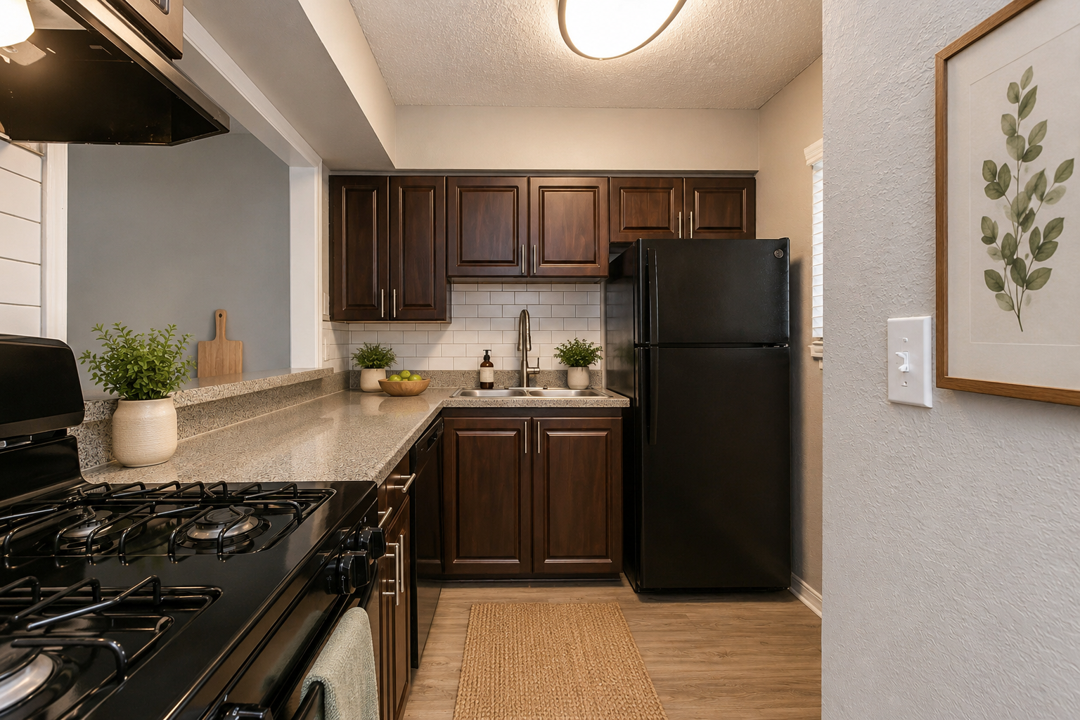 A modern kitchen featuring dark wooden cabinets and a granite countertop. There is a black refrigerator, a stove with a hood, and decorative plants on the counter. A light fixture hangs from the ceiling, and a framed botanical print is on the wall. The flooring is light wood.