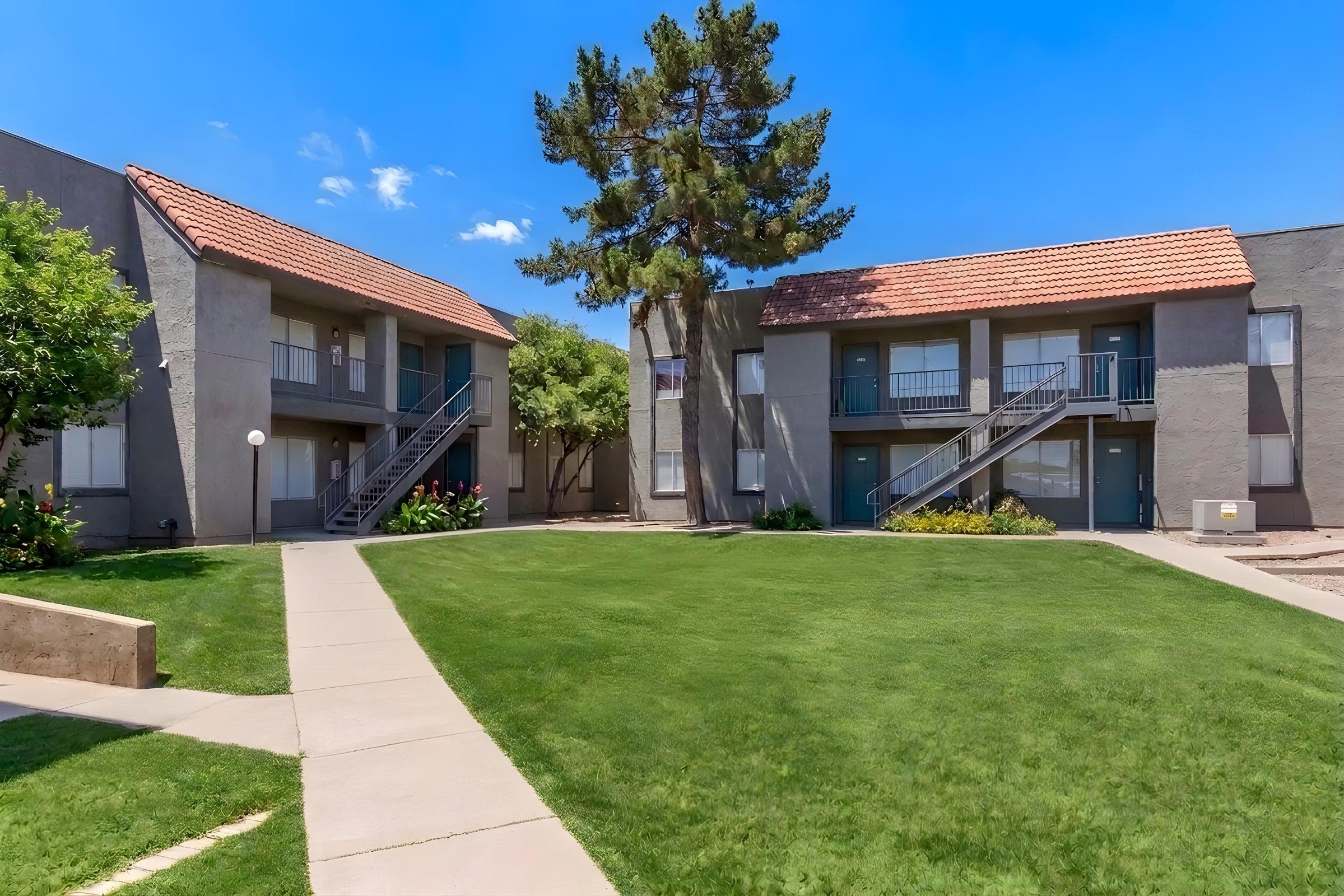 A peaceful apartment complex featuring two buildings with balconies, surrounded by green lawns and landscaped gardens. A pathway leads through the grass, with trees providing shade under a clear blue sky.
